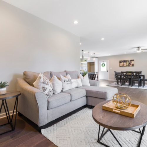 Living room with a light gray sectional sofa, decorative pillows, a round wooden coffee table with a geometric sculpture, a side table with a potted plant, and a dining area with a dark wood table and chairs in the background.