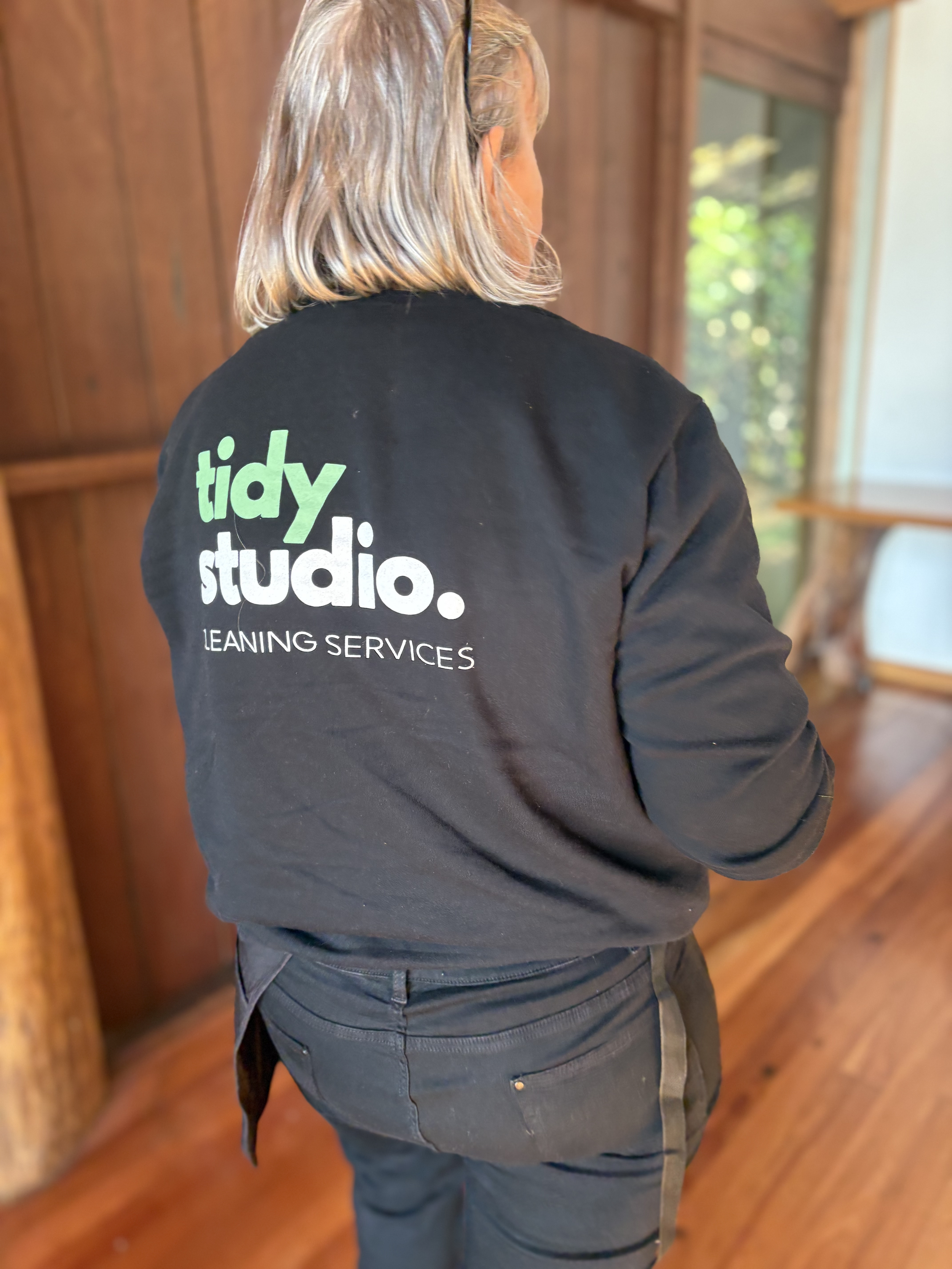 A person wearing a black jacket with the logo 'tidy studio' and text 'LEANING SERVICES' on the back, standing indoors with wooden walls and a window in the background.
