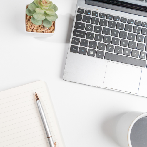 A workspace with a laptop keyboard, a small potted plant, a striped notebook with a pen, and part of a white mug on a white surface.