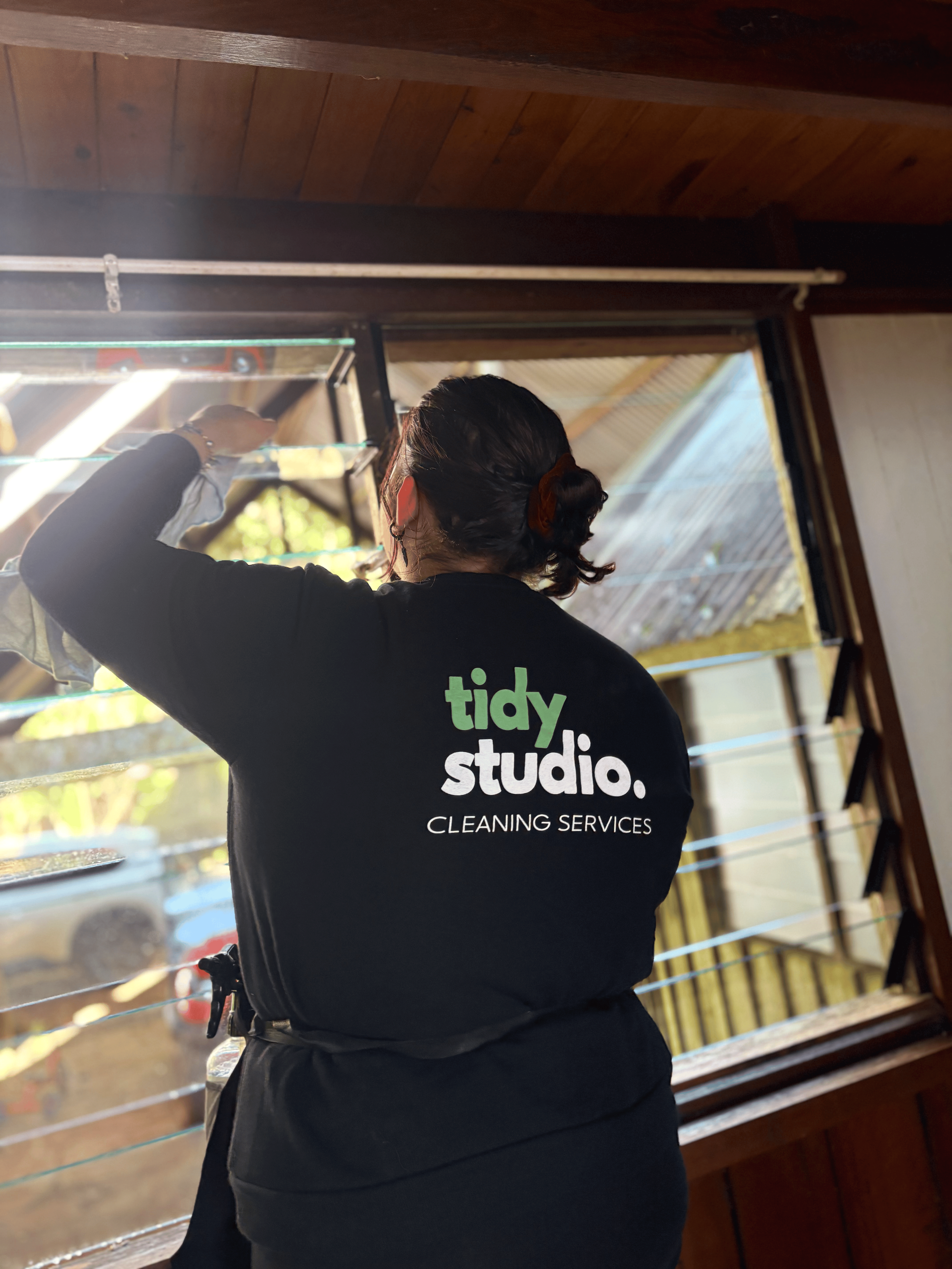 A person cleaning a window from inside a wooden building, wearing a black shirt with 'tidy studio. CLEANING SERVICES' printed on the back.