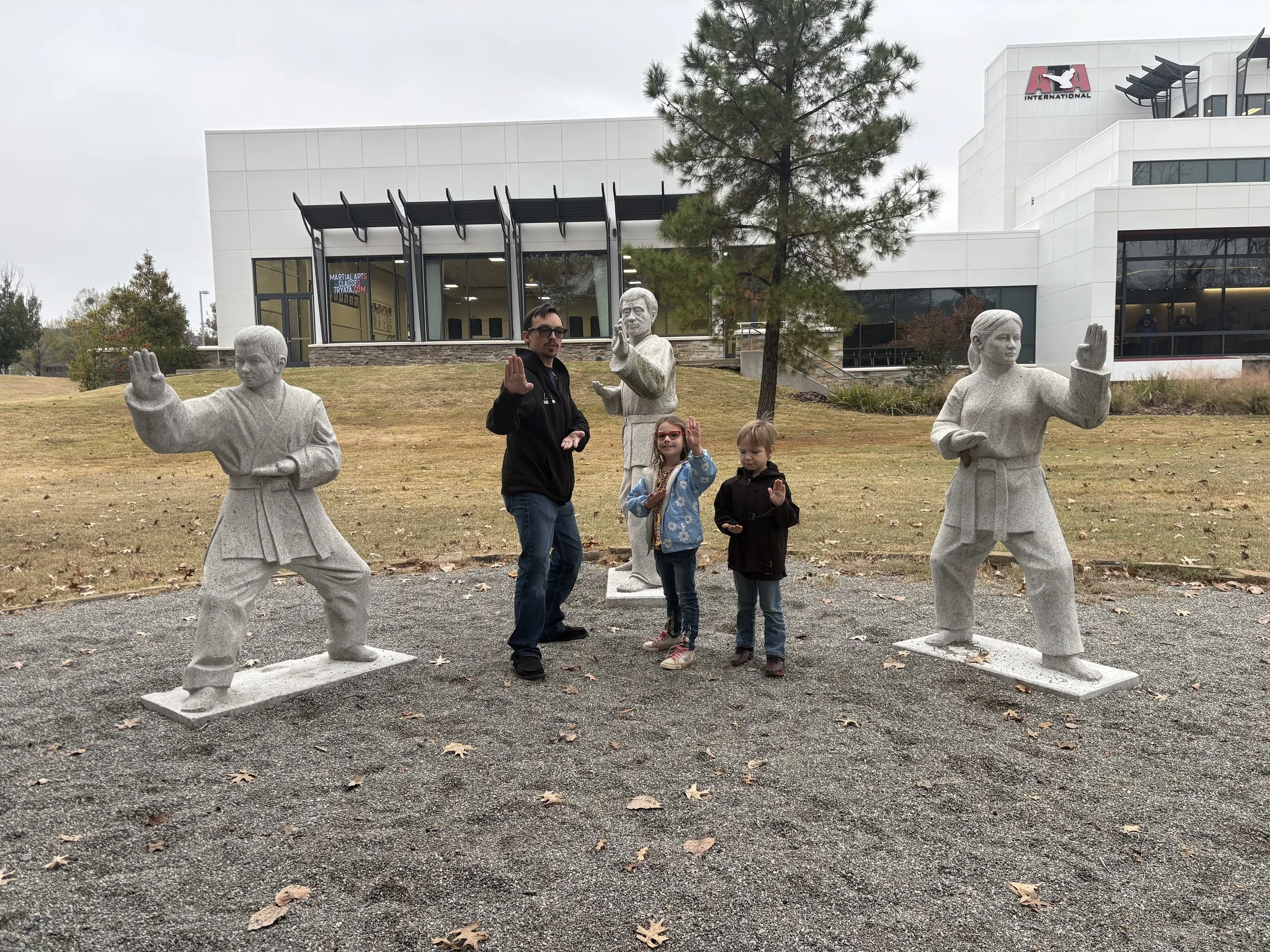 A man and two children posing with four martial arts statues outdoors in front of a modern building and trees.