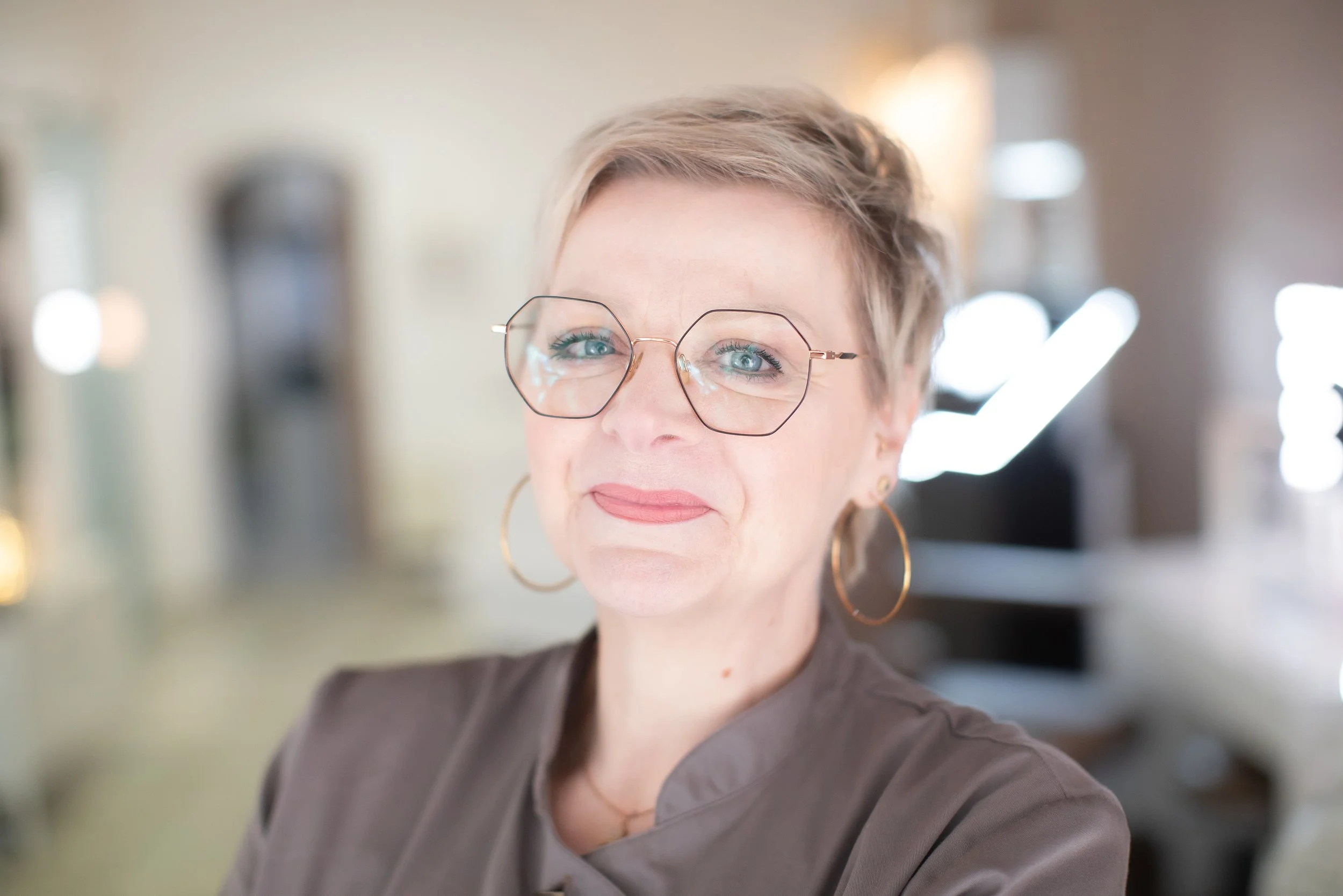 Eine Frau mit kurzen grauen Haaren, großen runden Brillen und goldenen Ohrringen, lächelnd in einem modernen Raum.