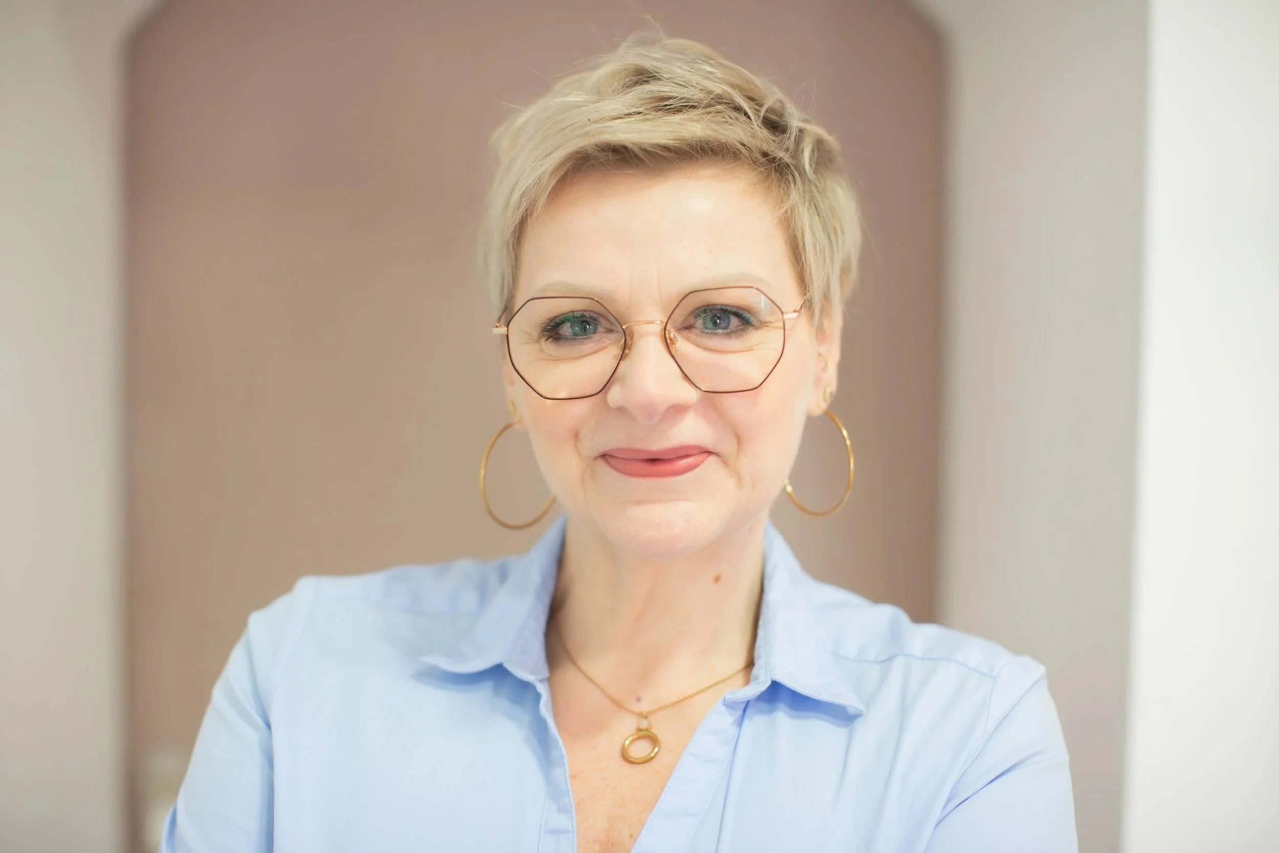 Nahaufnahme einer älteren Frau mit kurzen, blonden Haaren, großen runden Brillen, goldenen Ohrringen und einer goldenen Kette, trägt eine hellblaue Bluse, lächelt leicht, im Hintergrund unscharf beige Wände.