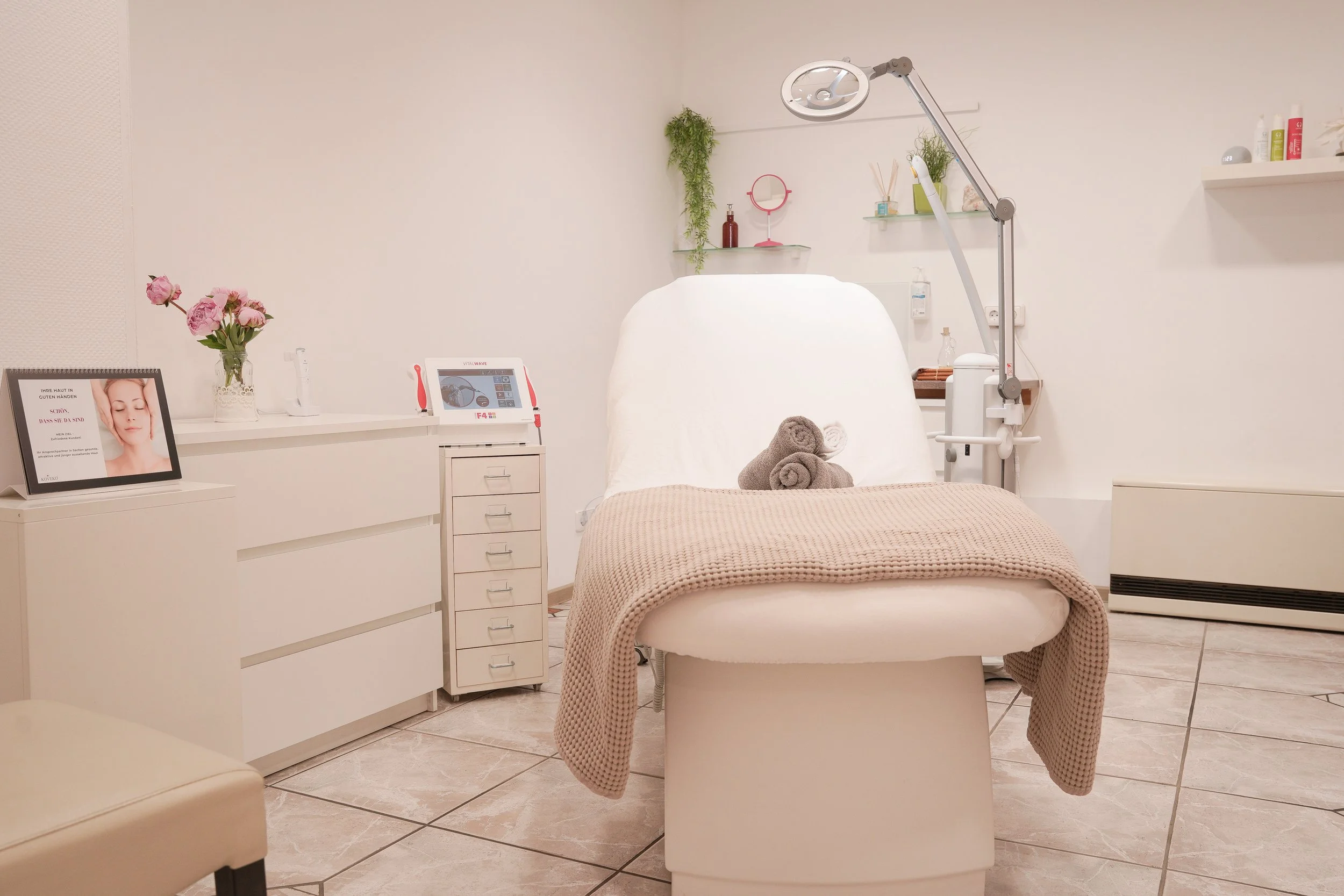Behandlungsstuhl in einer Kosmetik- oder Dermatologiesprechstunde, mit Handtüchern auf dem Stuhl, umgeben von medizinischen Geräten und Dekorationen in einem Behandlungsraum.