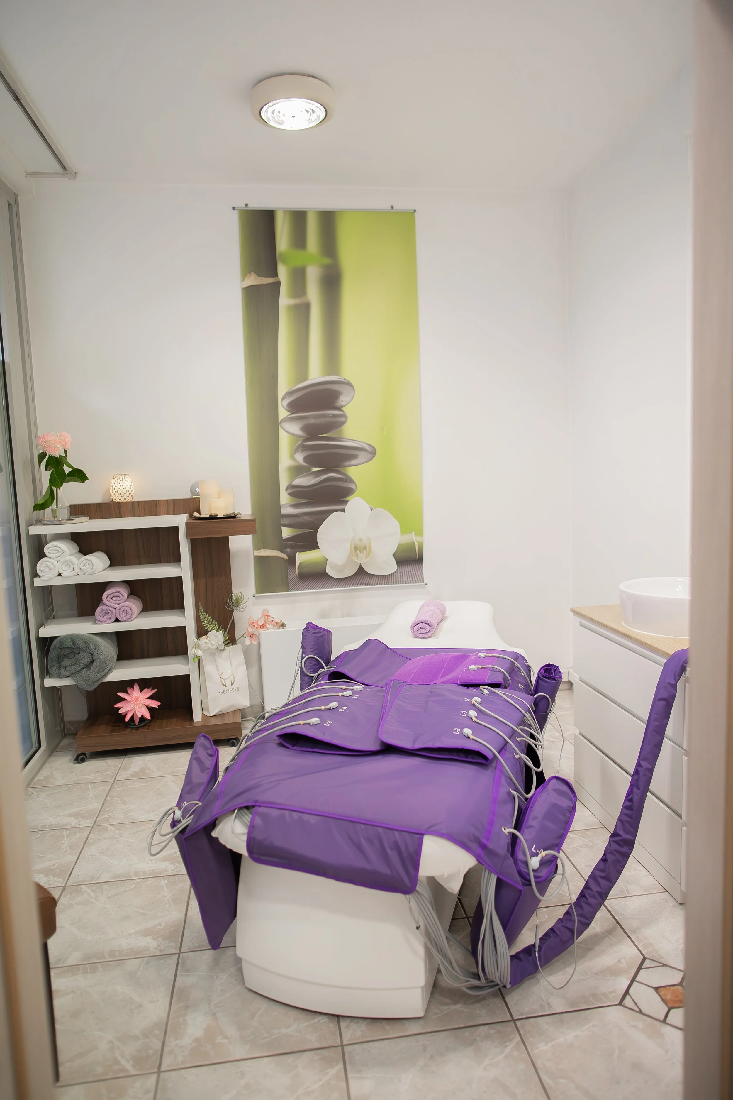 Massage- oder Wellnessraum mit Liege, die mit grüner und violetter Elektronik für Therapiezwecke bedeckt ist, neben einem Regal mit Handtüchern und Dekorationen, an der Wand ein Blumenbild.
