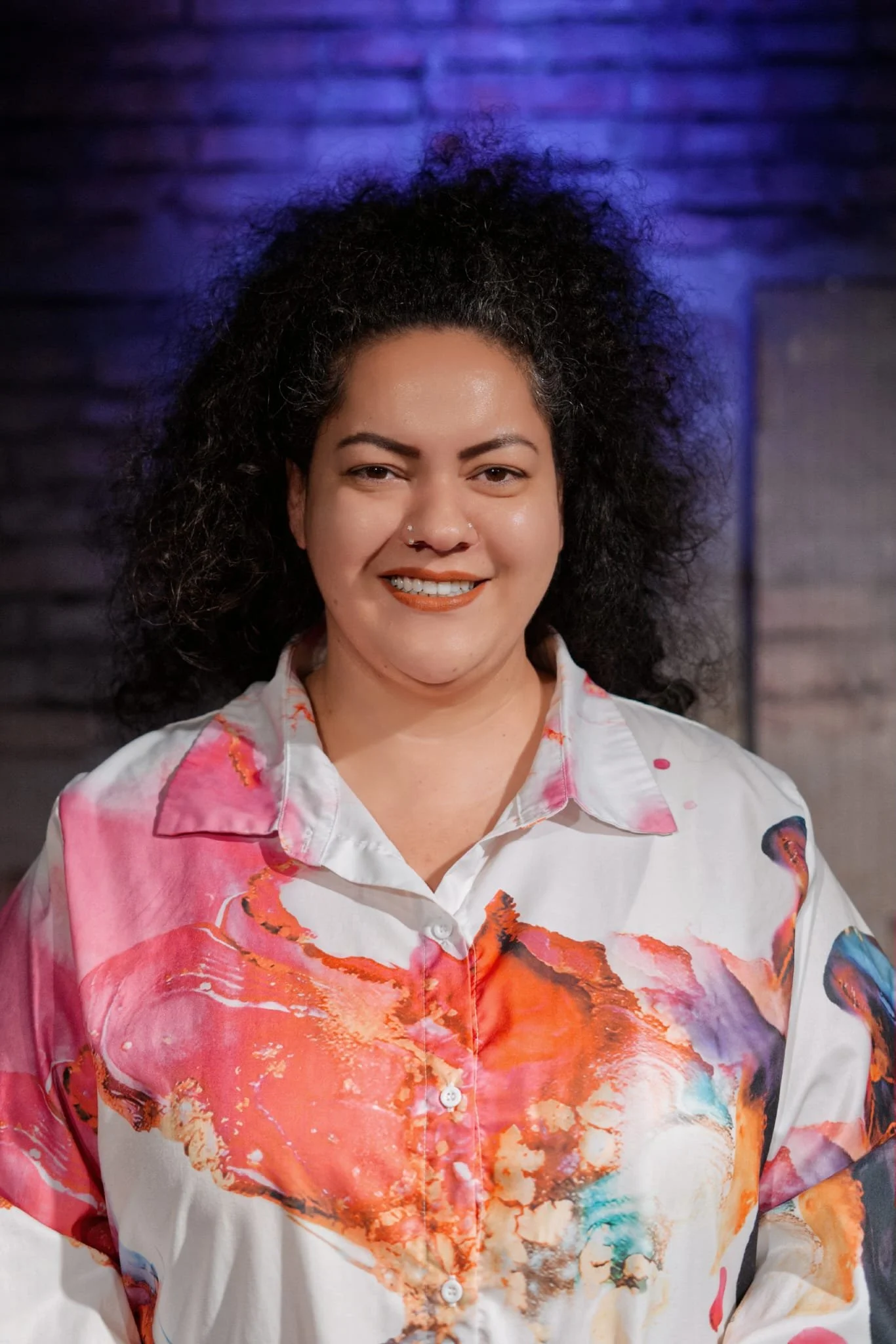 Portrait einer lachenden Frau mit dunklen, lockigen Haaren vor dunklem Hintergrund.