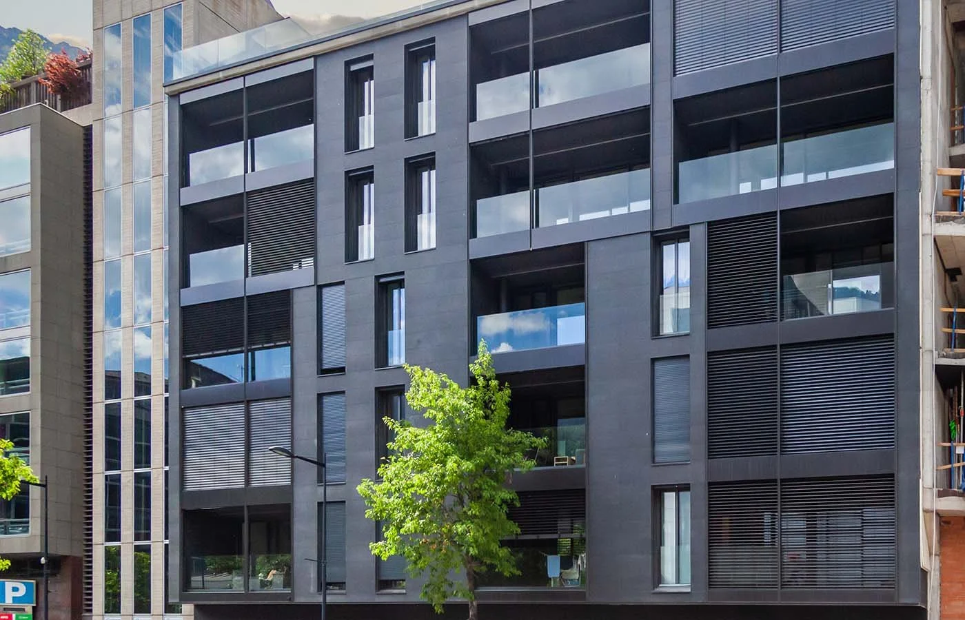 Edificio moderno con fachada de vidrio y ventanas grandes, un árbol verde en la acera en primer plano.