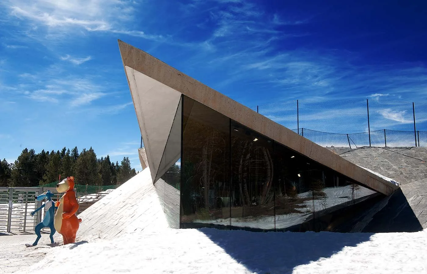 Edificio moderno con forma angular y grandes ventanas de vidrio, rodeado de nieve, con estatuas de personajes de dibujos animados de Tom y Jerry a la izquierda, y un cielo azul con nubes delgadas arriba.