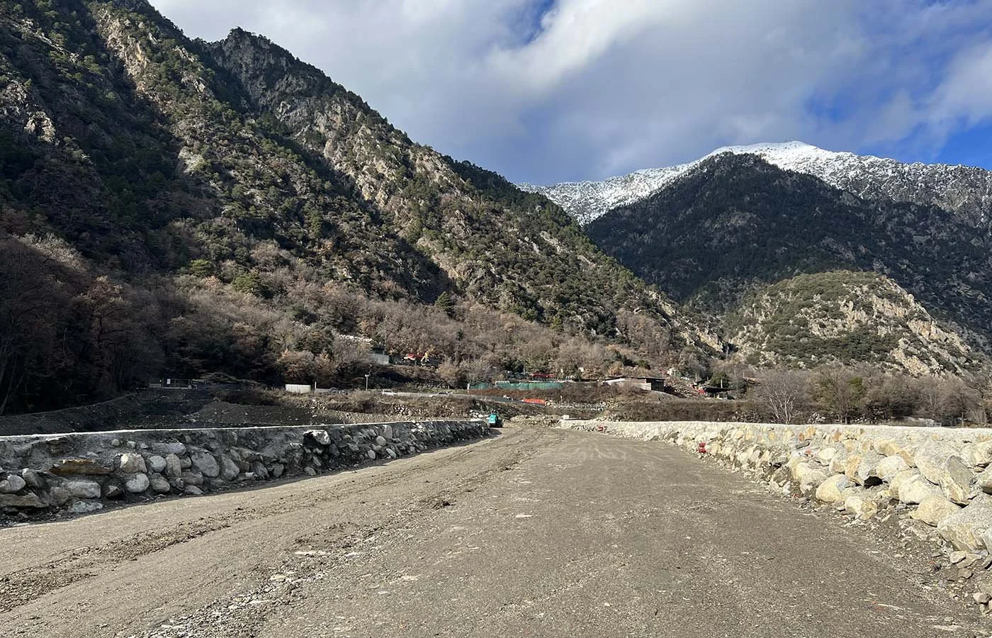Camino de tierra en un valle rodeado de montañas con algunas casas pequeñas y nieve en las cumbres al fondo.