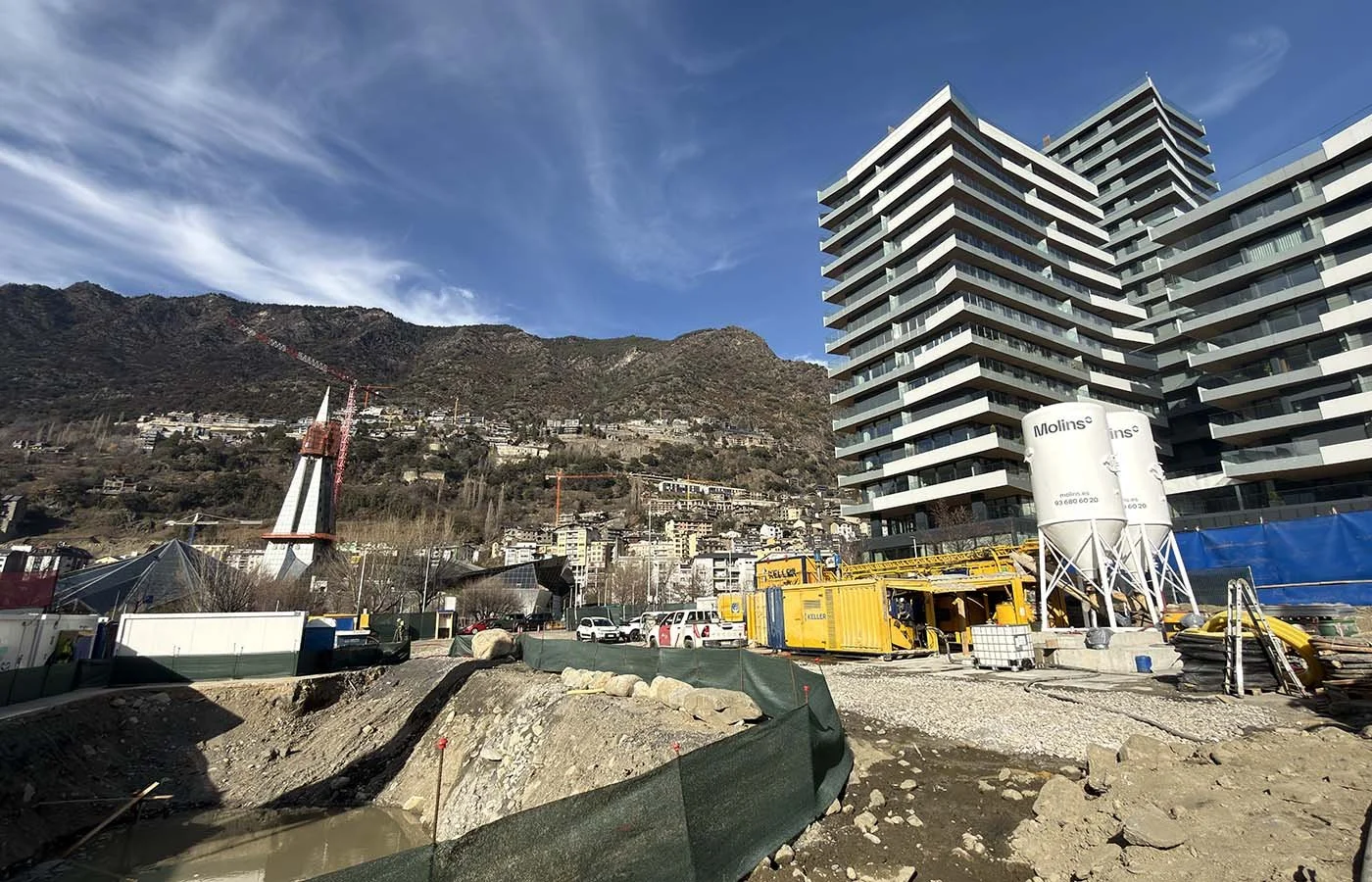 Sitio de construcción con equipo y maquinaria frente a edificios altos y montañas bajo cielo despejado.
