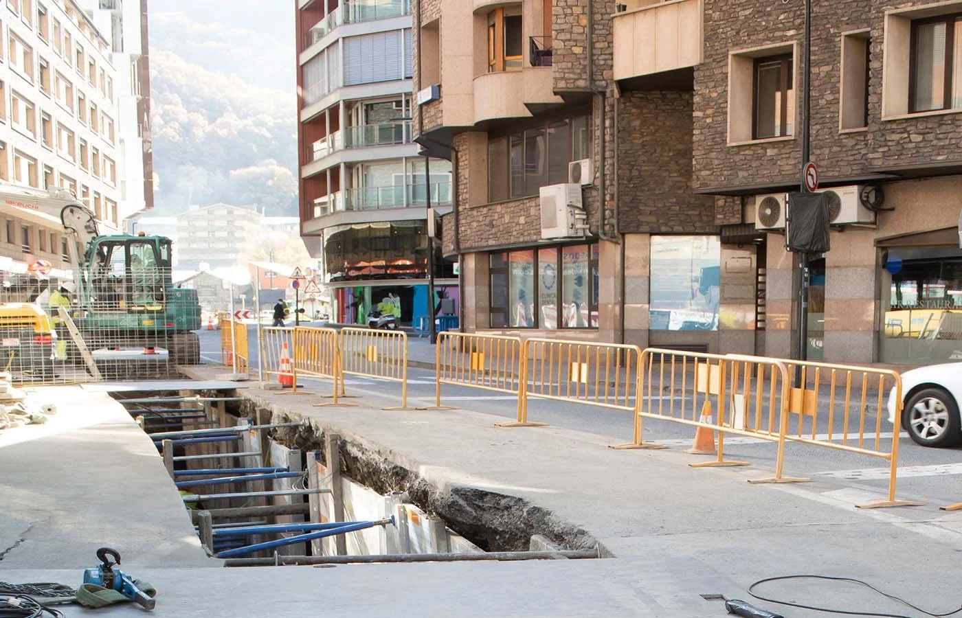 Construcción en la calle con barreras amarillas, una excavadora y un hueco en el suelo, en medio de edificios residenciales y negocios.