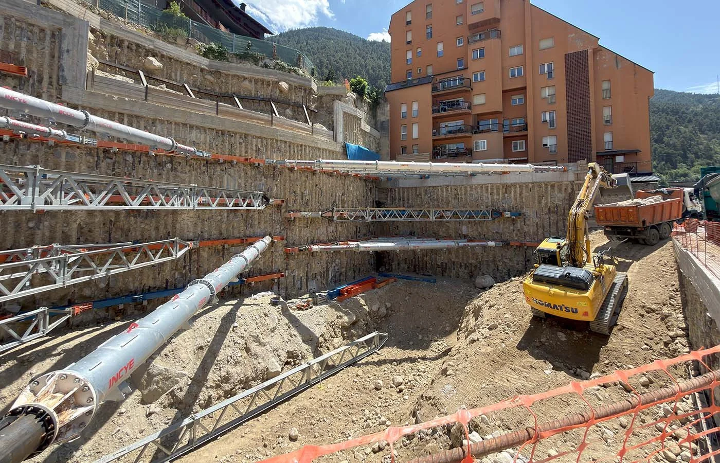 Obra en construcción con excavadora amarilla y tuberías instaladas en una ladera con montañas de fondo bajo cielo azul.