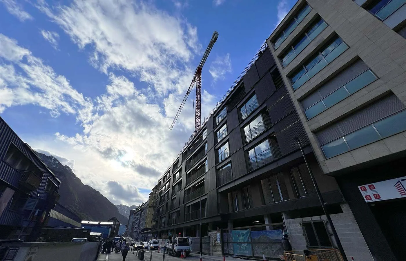 Edificio en construcción con grúa en un área urbana rodeada de montañas y cielo con nubes.