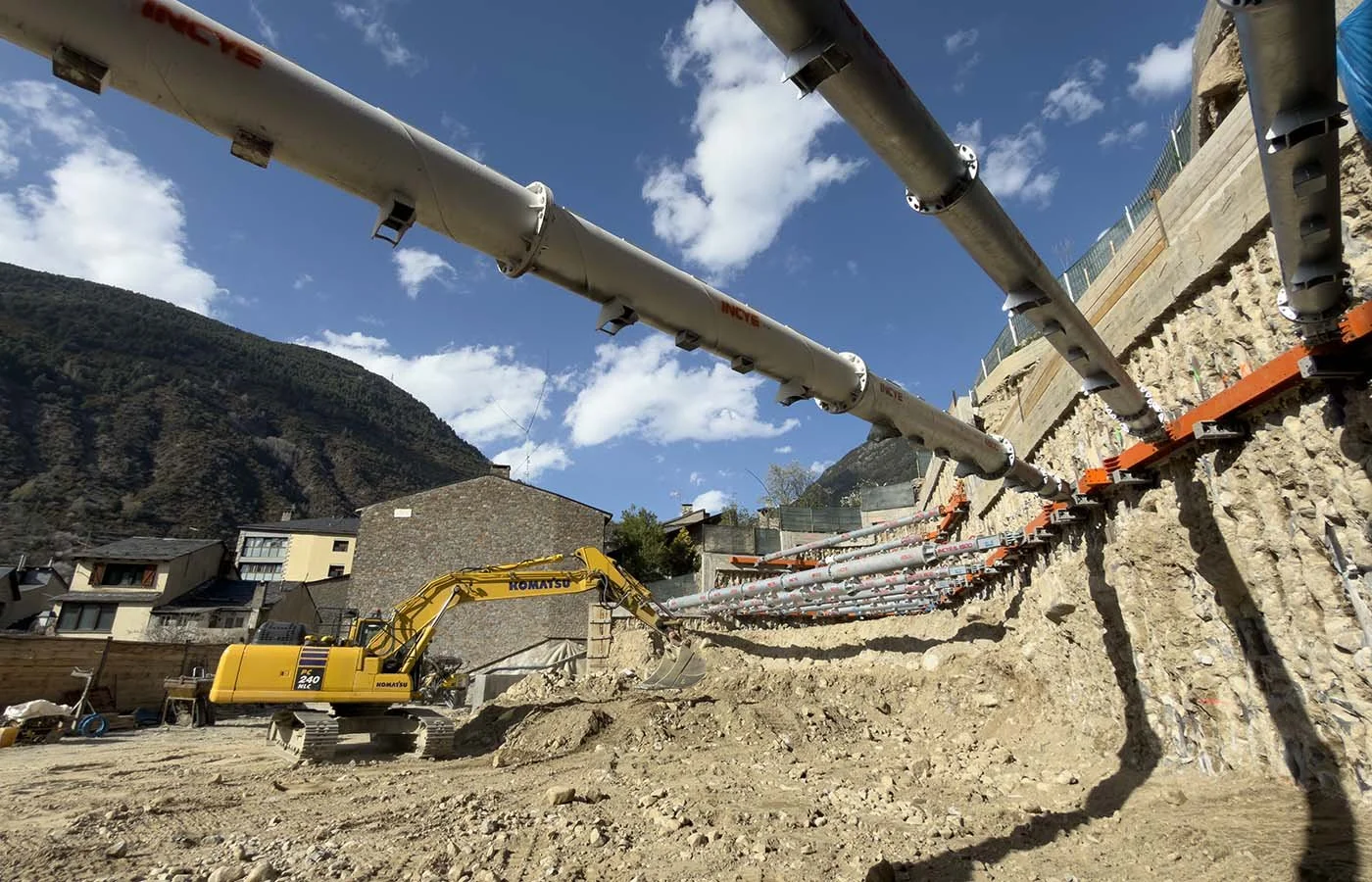 Obra en construcción con excavadora amarilla y tuberías instaladas en una ladera con montañas de fondo bajo cielo azul.