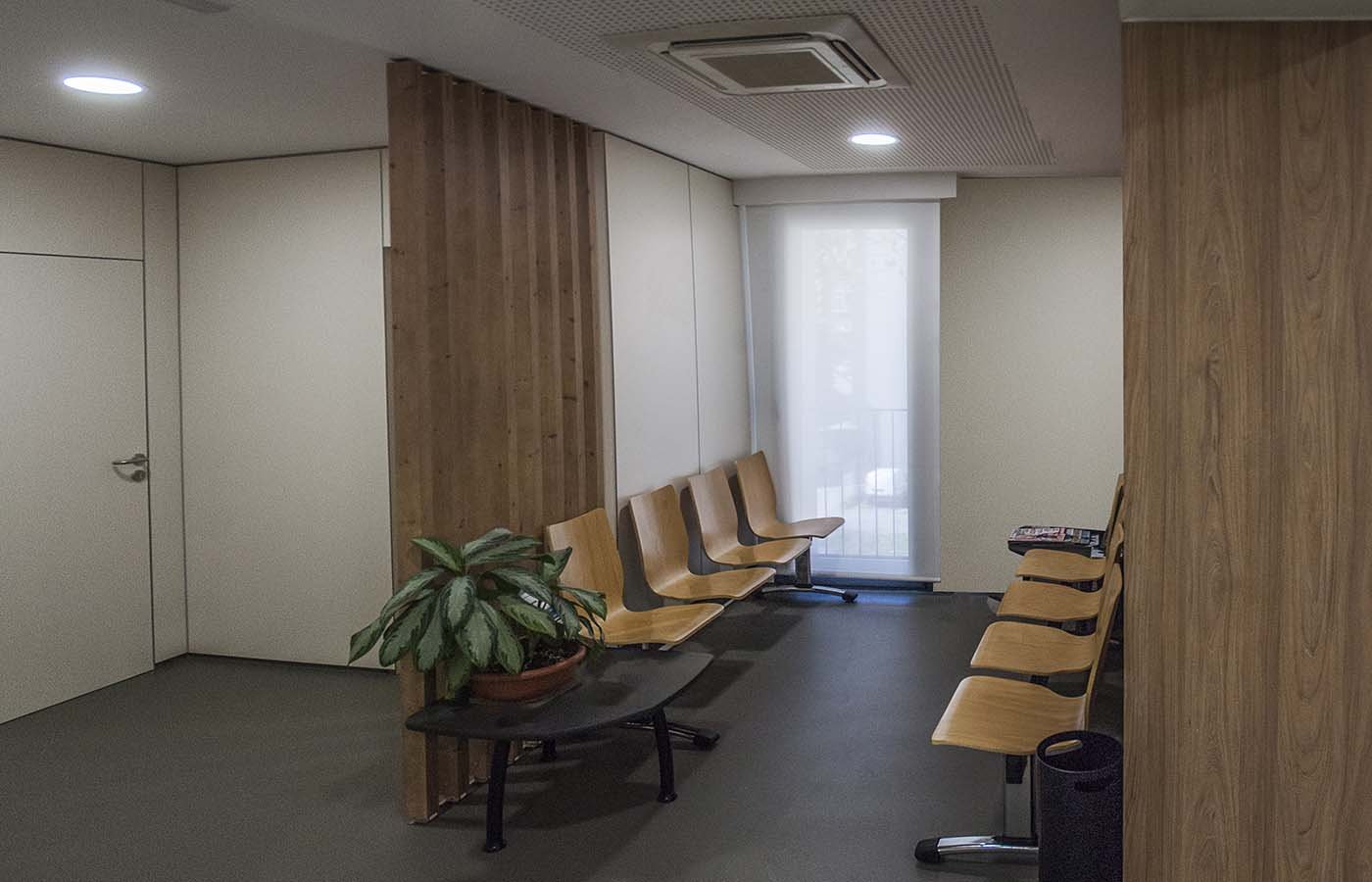 Sala de espera con sillas de madera, planta en maceta, ventanas con cortinas, pared de madera y paredes blancas.