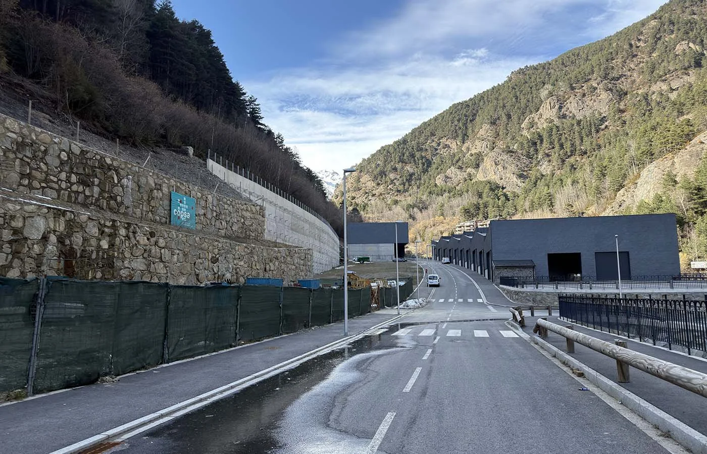 Camino en una zona montañosa con construcciones y vehículos y un muro de contención de roca y hormigón a la izquierda.
