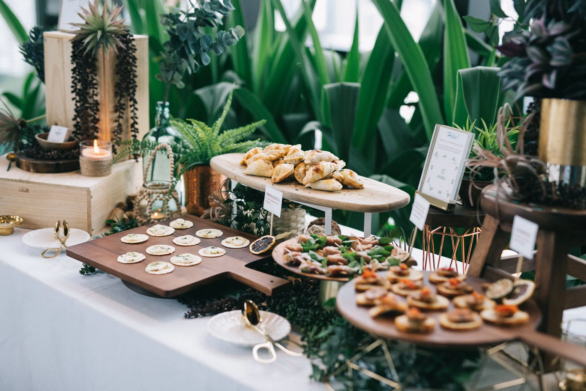 Appetizer table with various finger foods, greenery, candles, and decorative items, set on a white tablecloth with lush green plants in the background.
