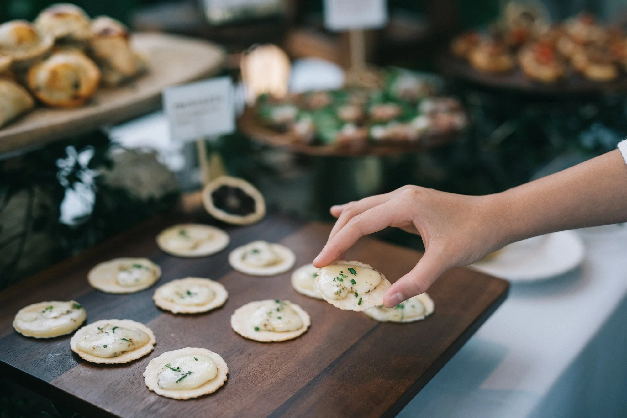 Hand placing a canapé with cheese and herbs on a wooden tray, with more appetizers and food trays in the background.