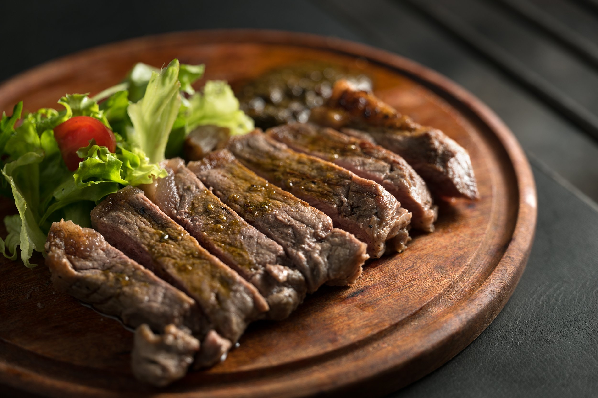 Sliced grilled steak with green salad and cherry tomato on a wooden serving board.