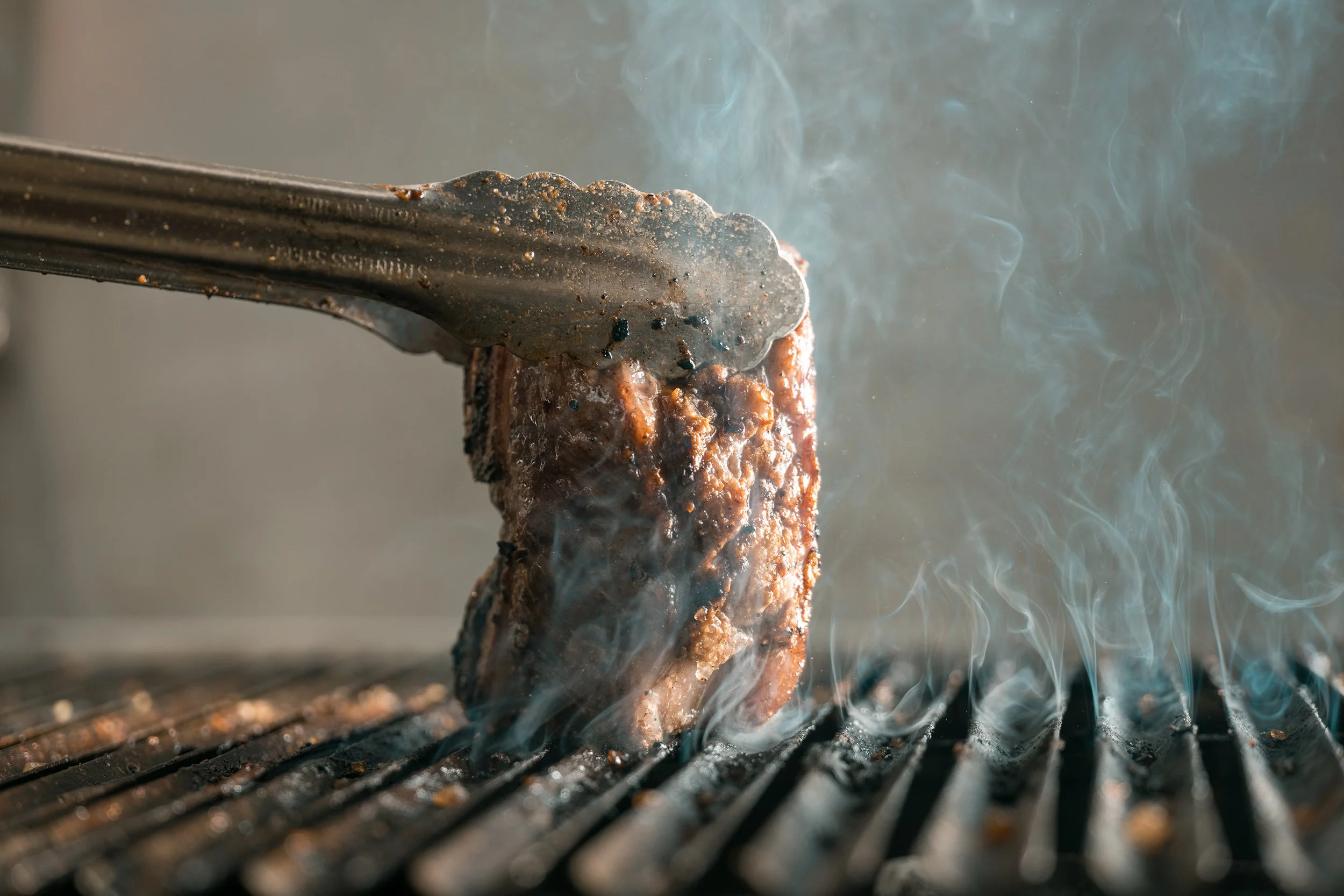A piece of steak being cooked on a grill with tongs holding it, steam rising from the meat.