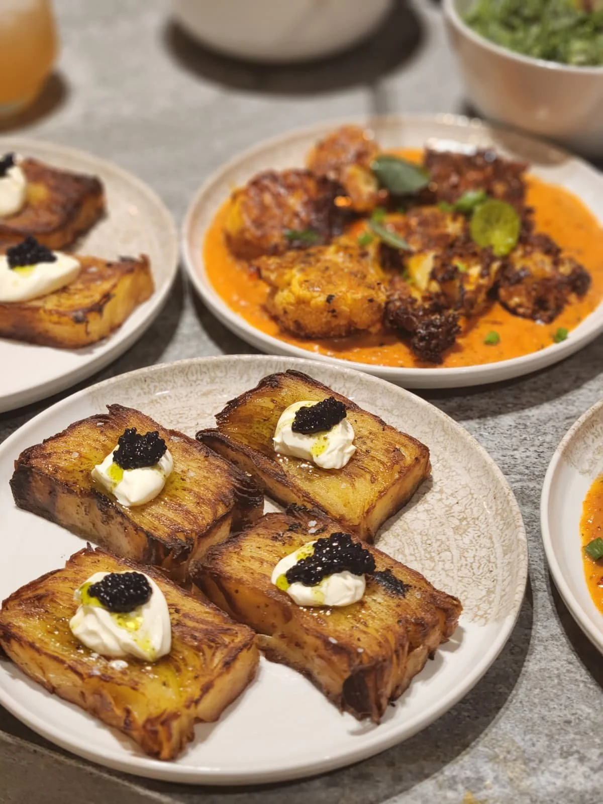 Close-up of a white plate with four grilled pieces of plantains topped with dollops of cream and caviar. In the background, there are other plates with various dishes, including a spicy meat dish in an orange sauce garnished with green herbs.