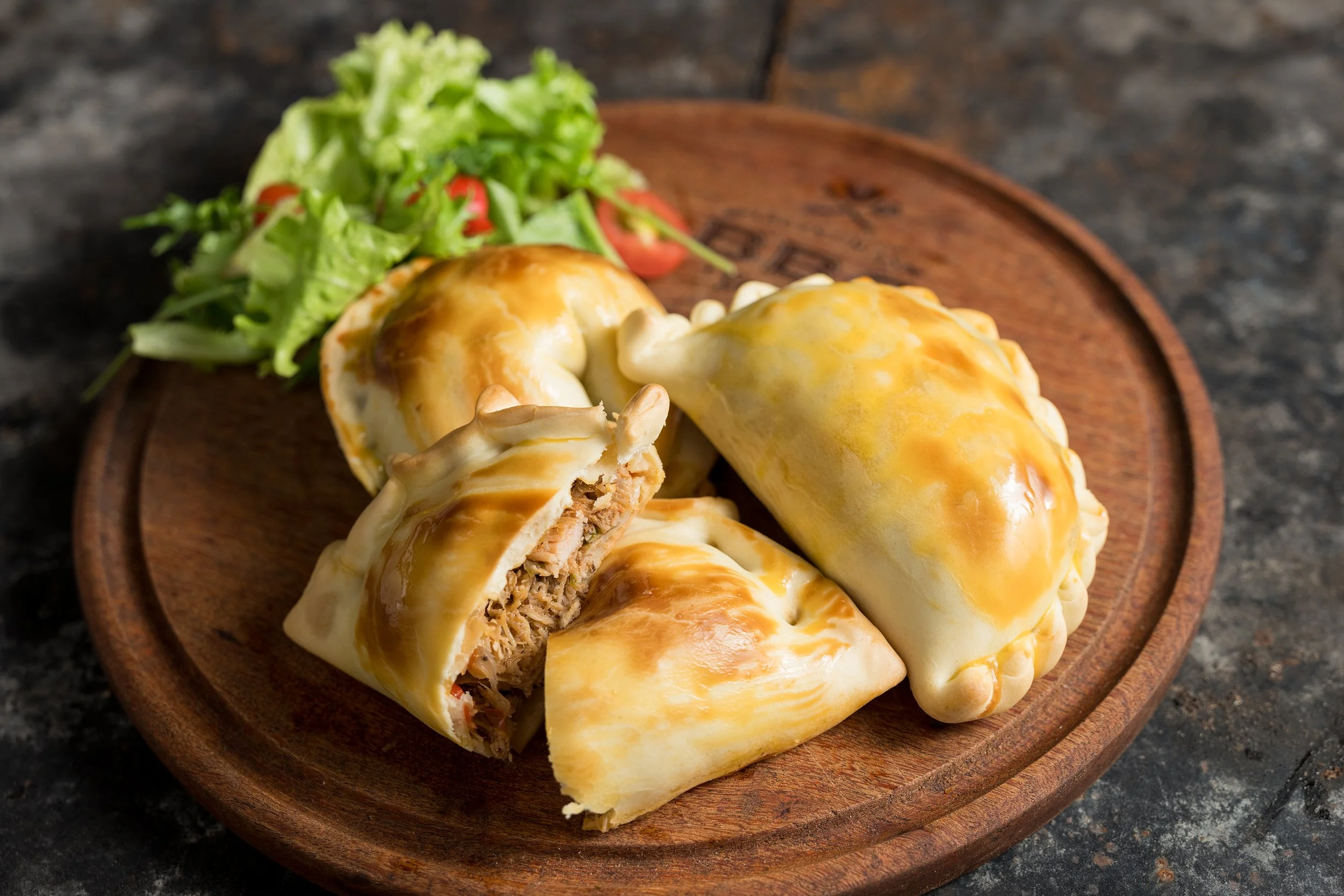 Four chicken and cheese empanadas on a wooden plate with a side of green salad.