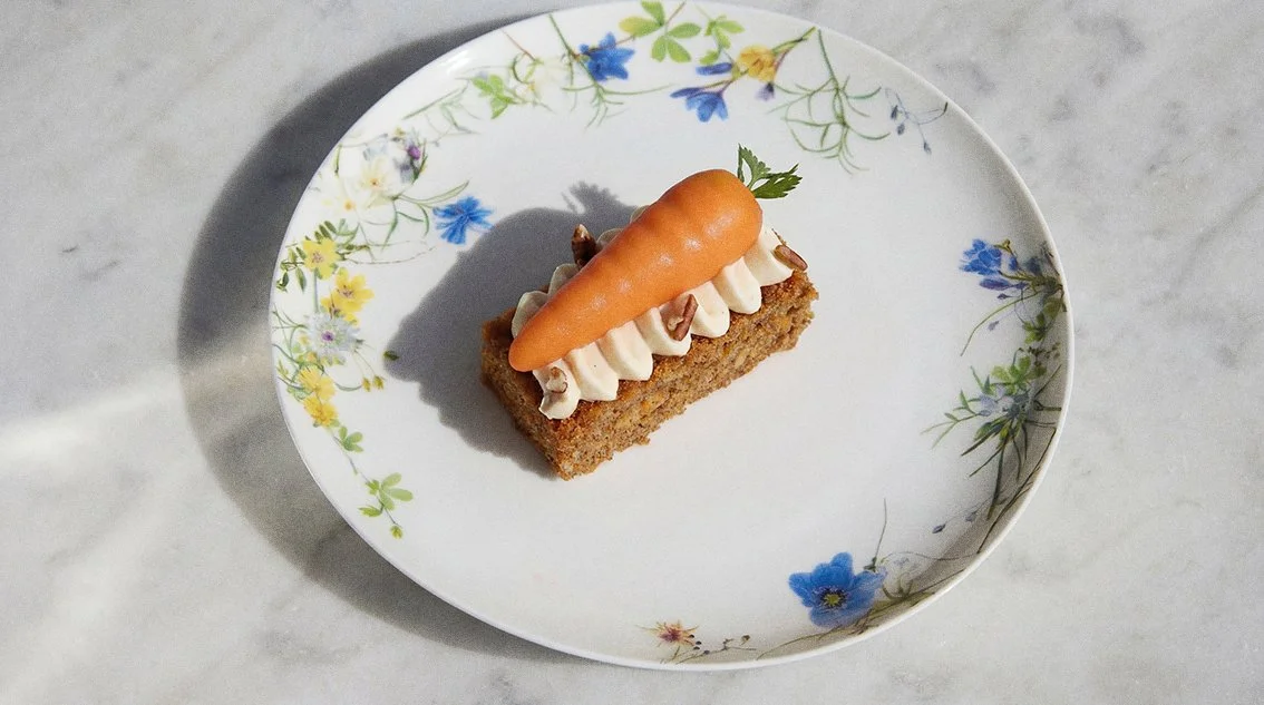 Ein Dessert auf einem weißen Teller mit Blumenmuster, bestehend aus einem rechteckigen Kuchenstück, gefüllt mit Sahne und Nüssen, garniert mit einem kleinen Karottenmodell.