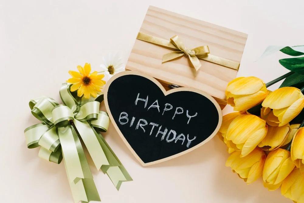 Blumen, eine Geschenkbox mit Goldband, eine Herz-Schiefertafel mit 'Happy Birthday' und gelbe Tulpen.