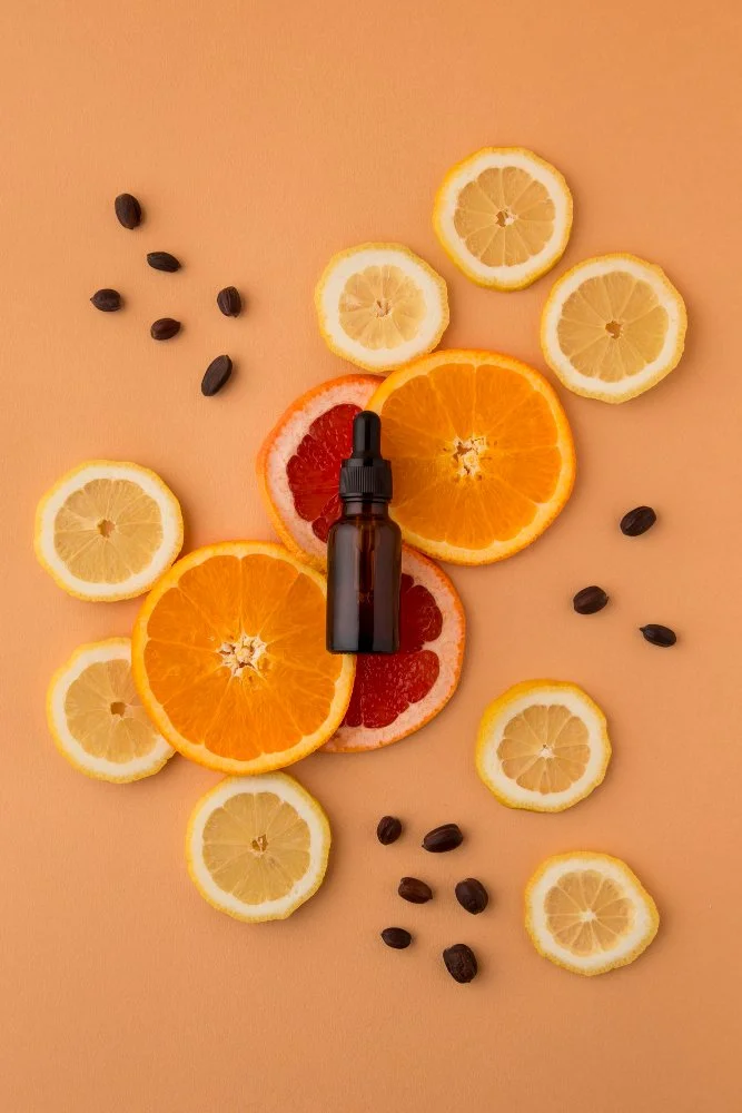 Schnittfrüchte von Zitrone, Orange und Grapefruit, umgeben von schwarzen Samen und einer braunen Flasche mit Pipette auf einem peachfarbenen Hintergrund.