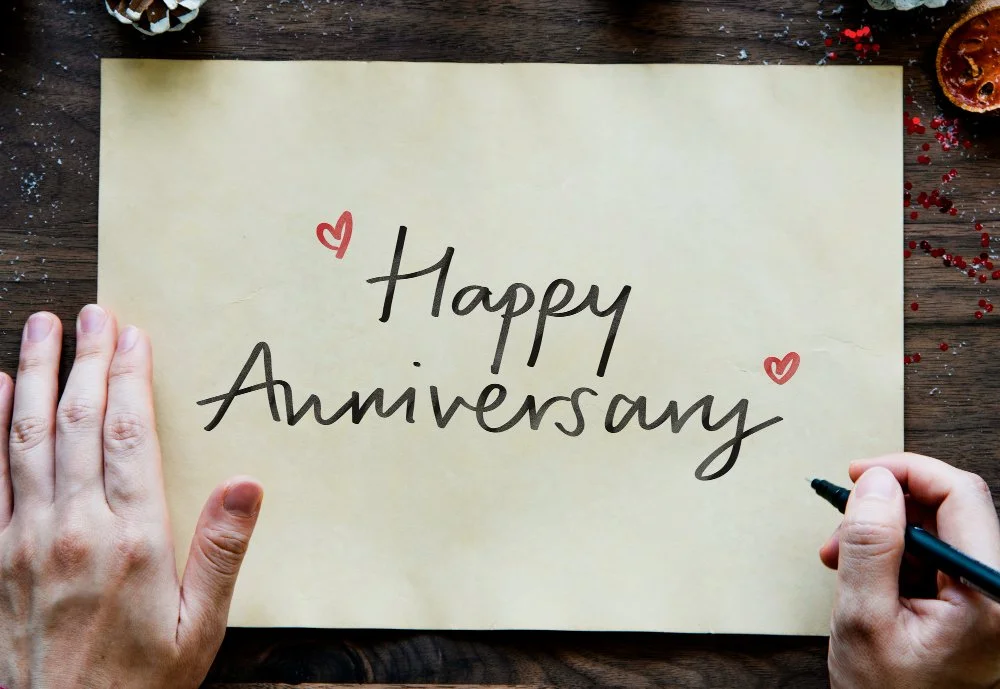 Eine Person schreibt die Worte »Happy Anniversary« auf ein gelbes Blatt Papier, begleitet von kleinen roten Herzzeichnungen, auf einem dunklen Holztisch mit einigen Dekorationsstücken und Konfetti.