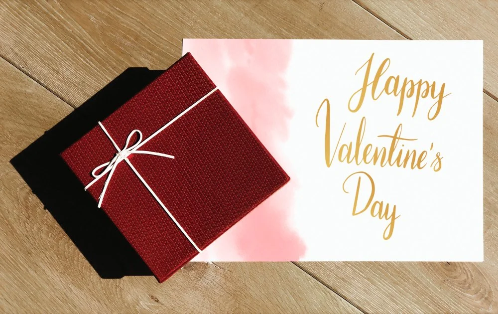 Ein Geschenk in rotem Papier mit weißer Schnur auf einem Holztisch neben einer Grußkarte, die 'Happy Valentine's Day' auf Englisch schreibt.