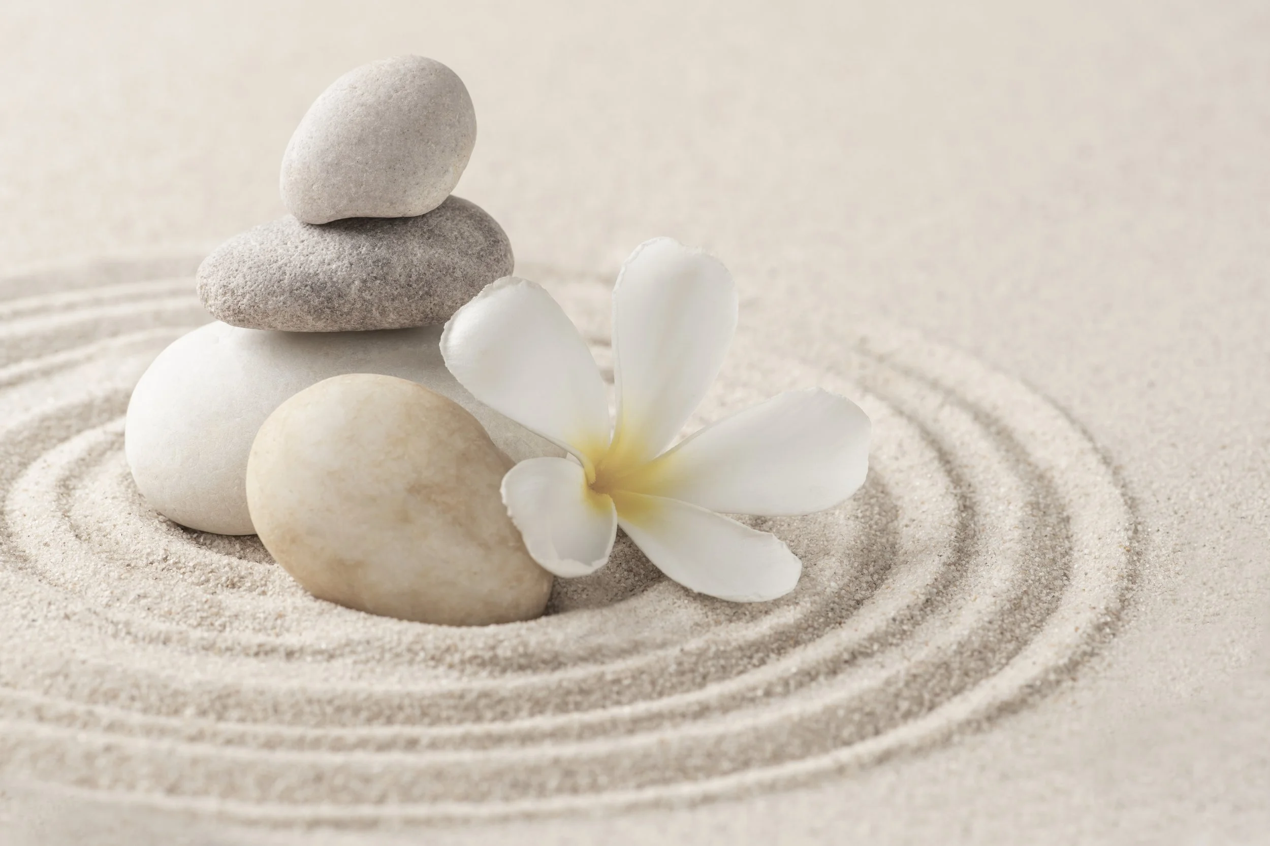 Sand, ideal for relaxation, with white stones, a white flower, and a balanced stack of three stones on top.