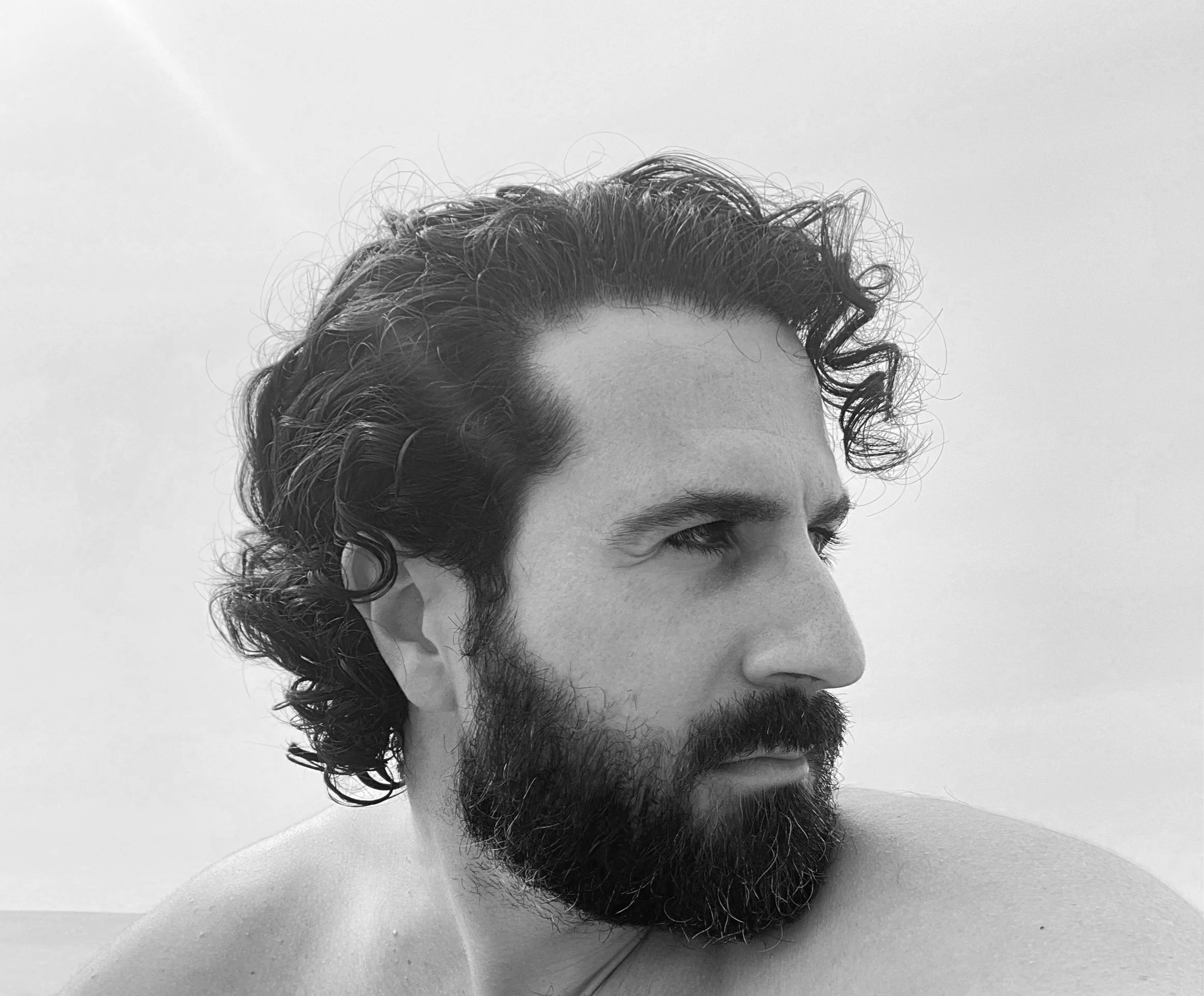 Black and white close-up image of a man with curly hair and a beard, looking to the right.
