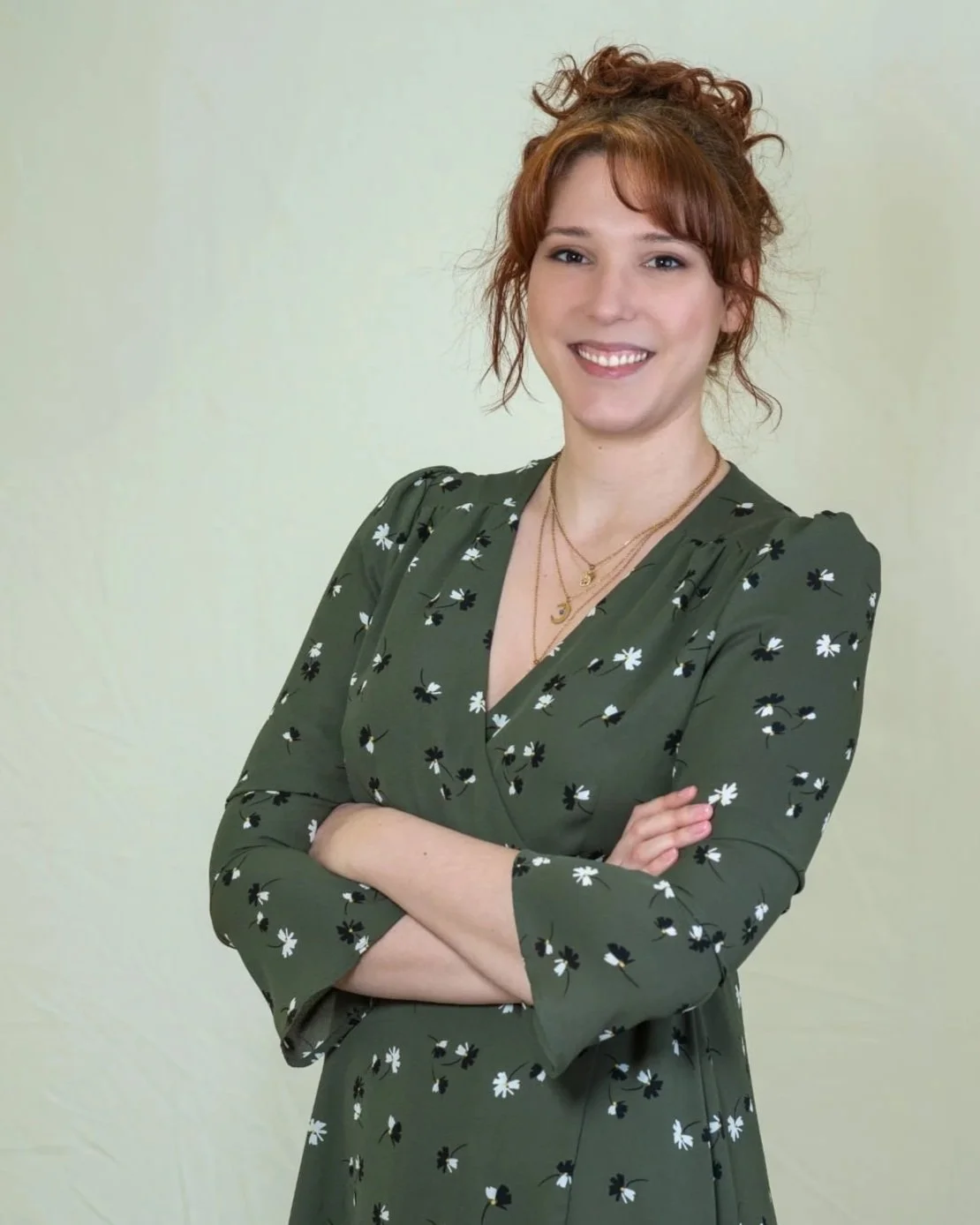 A young woman with red curly hair smiling, wearing a green floral dress with layered necklaces, standing against a light plain background.