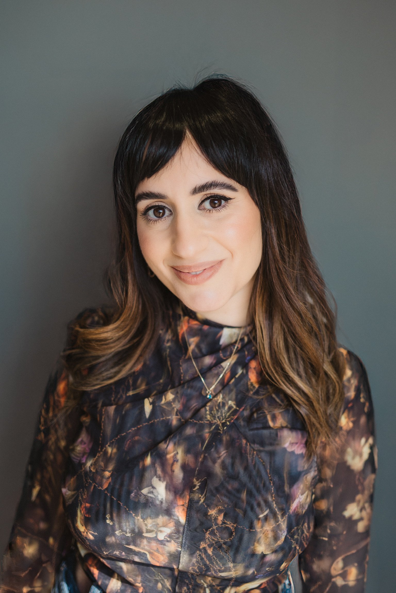 A woman with shoulder-length brown hair and bangs, wearing a dark floral patterned blouse, smiling against a plain gray background.