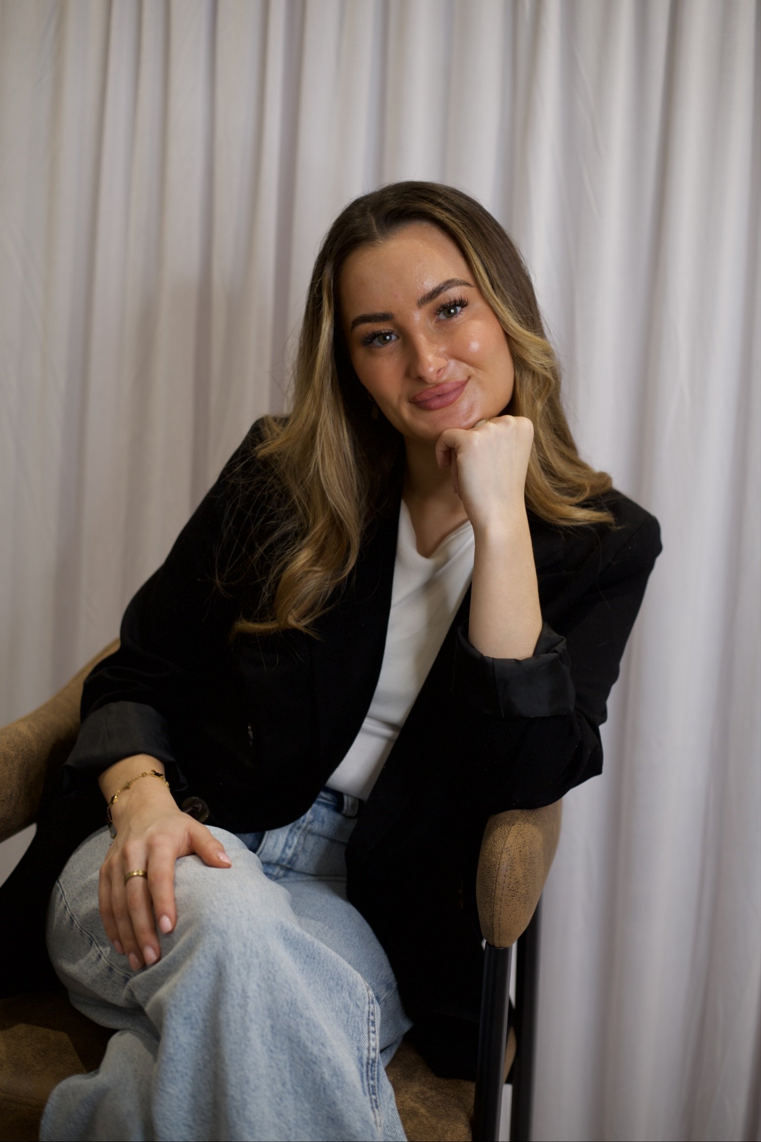 A young woman with long, wavy hair and light makeup sitting in a beige armchair in front of a white curtain, resting her chin on her fist and looking at the camera with a slight smile.