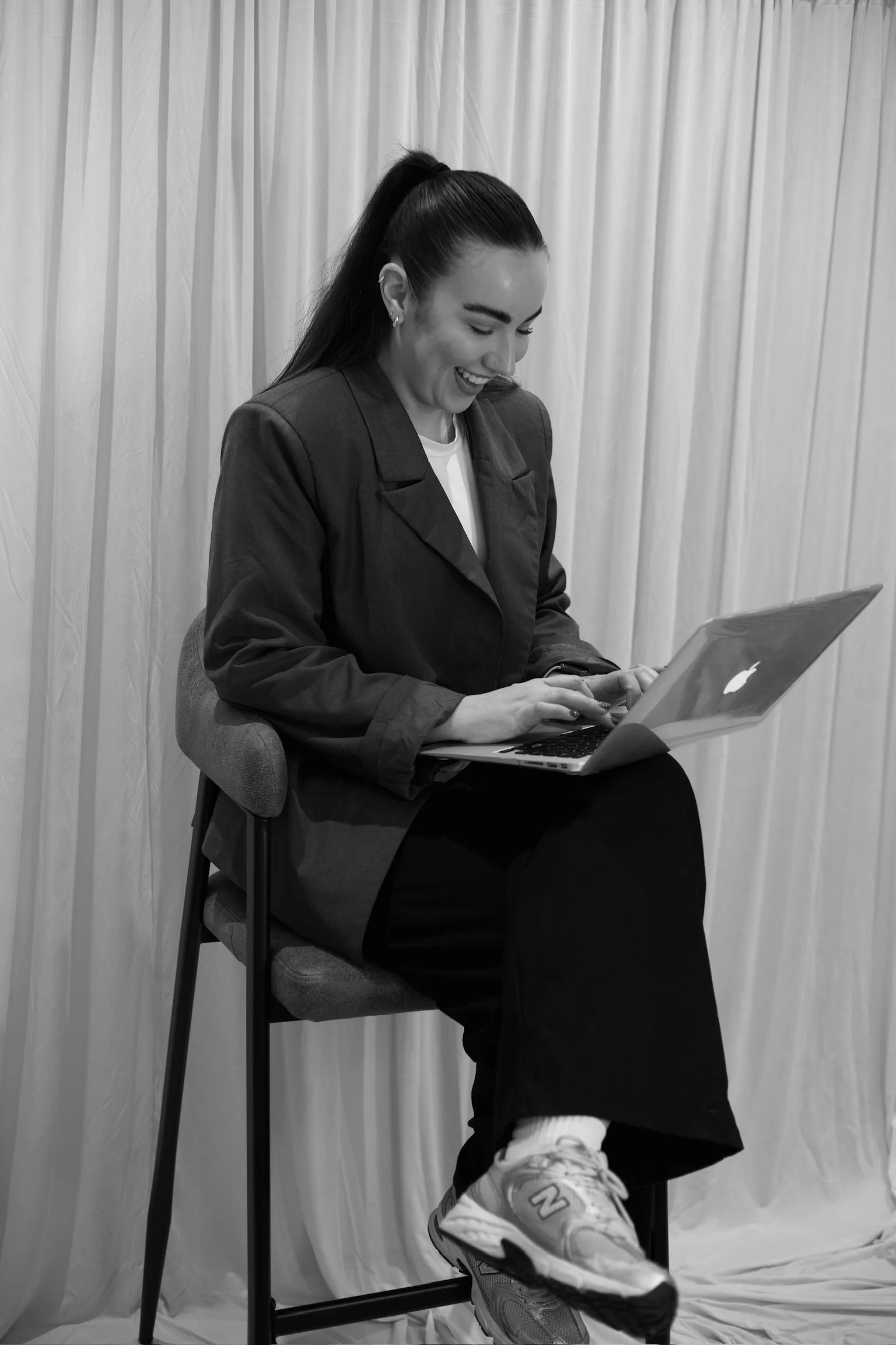 A woman sitting on a chair with a laptop on her lap, smiling, and wearing casual clothing and sneakers, with a light-colored curtain as the background.