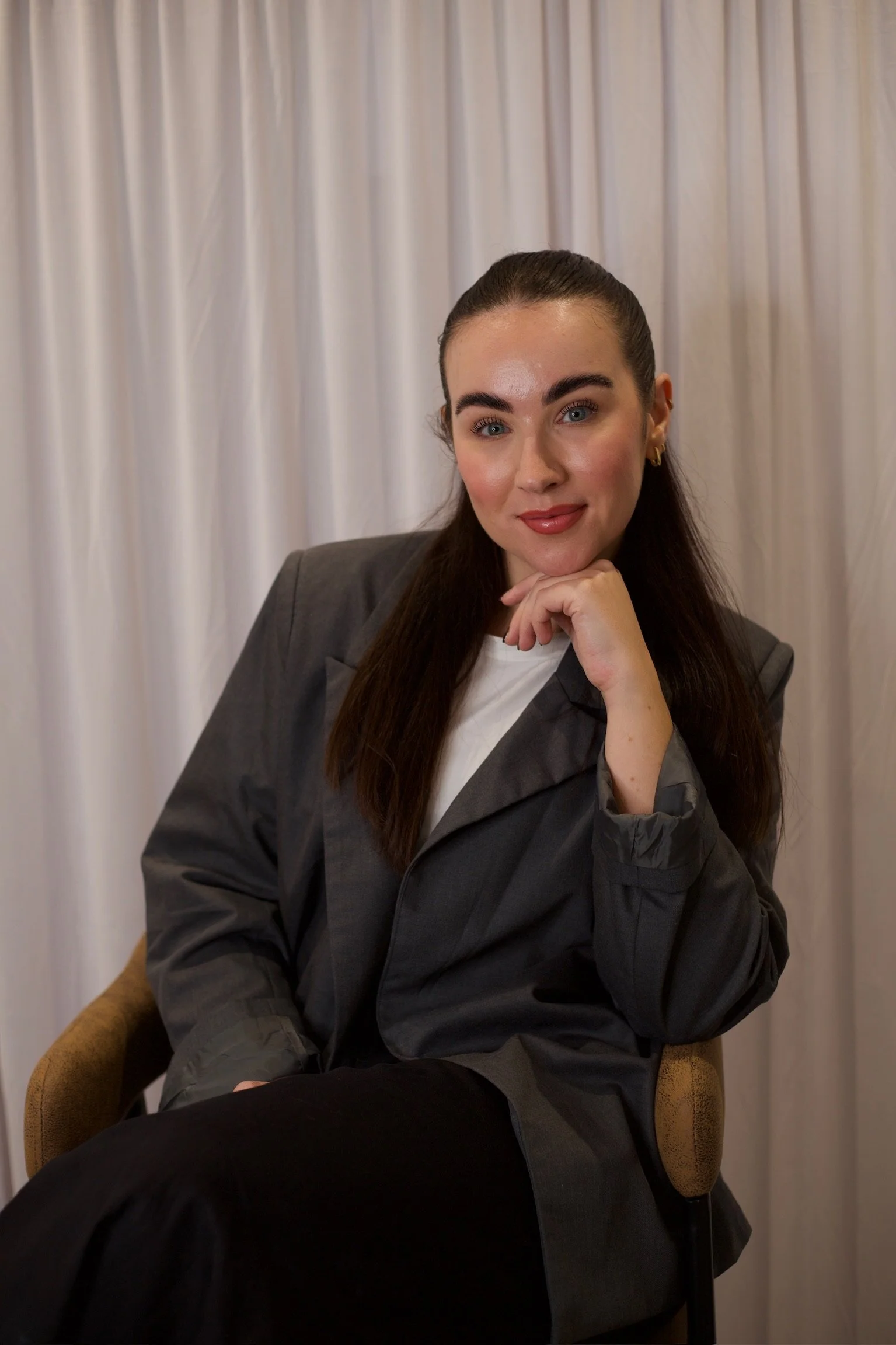 A woman with long dark brown hair, wearing a gray blazer and white top, sitting on a chair in front of a white curtain, smiling with her hand resting on her chin.