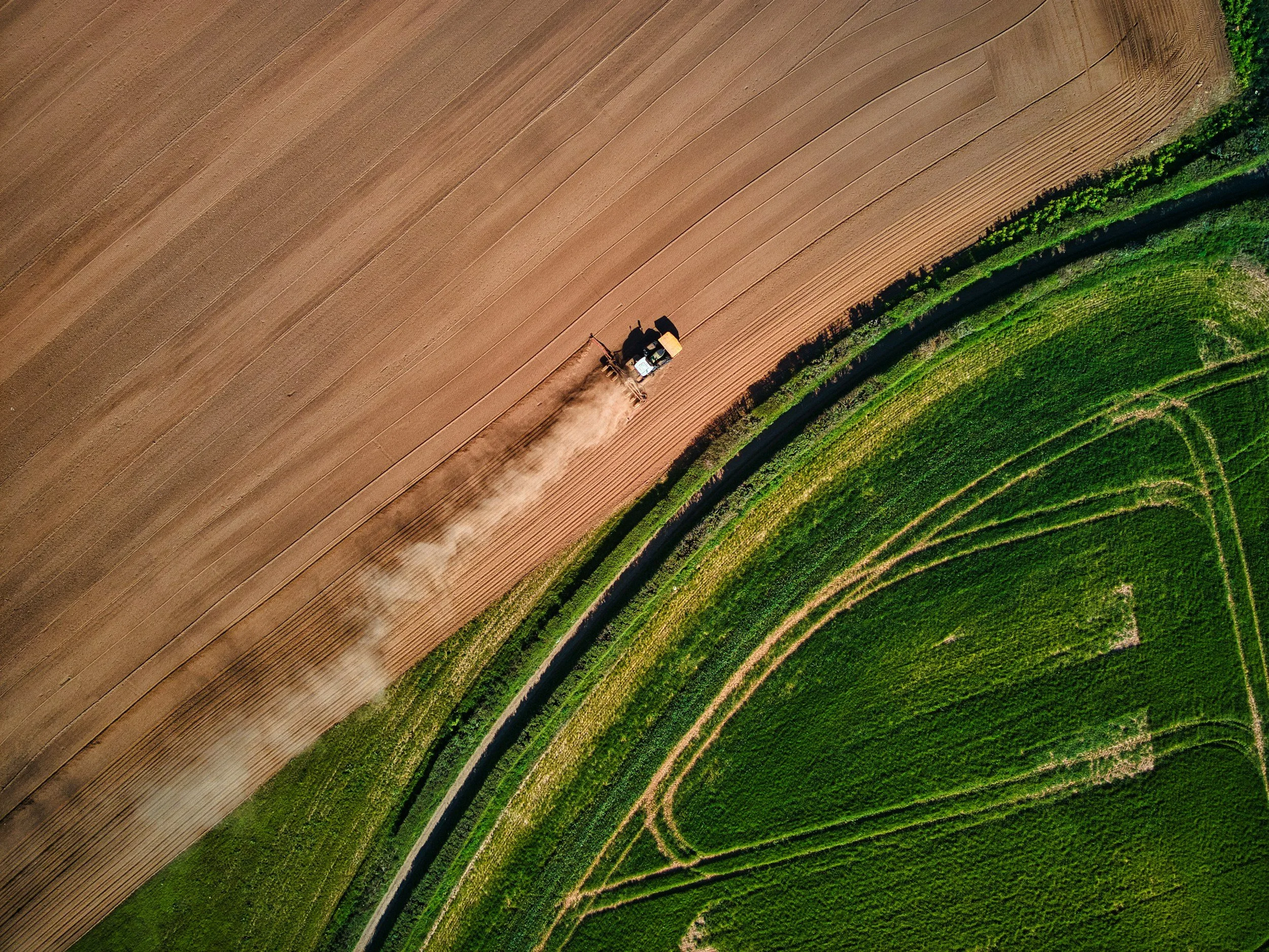 Eine Drohnenaufnahme zeigt landwirtschaftliche Felder, bei denen eine Maschine den Boden bearbeitet, Staub aufwirbelt. Die Felder sind in verschiedenen Grüntönen und Braunfarben gehalten.