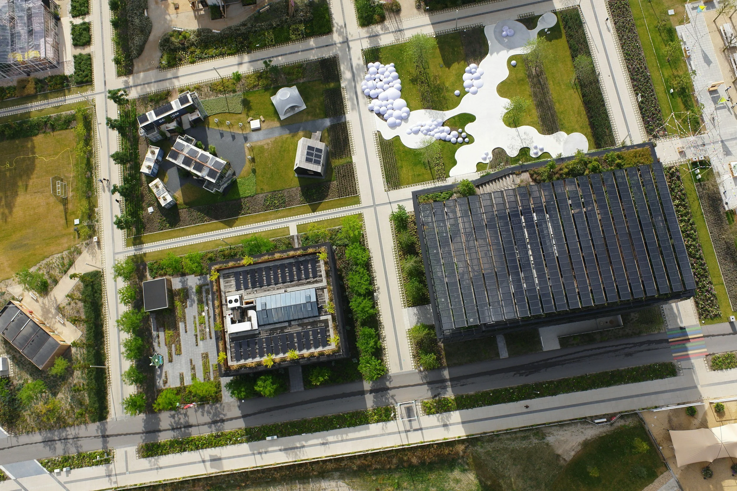 Luftaufnahme eines Stadtparks mit weißen Skulpturen, Gebäuden mit Solarpanel-Dächern und Wegen, umgeben von urbanen Gebäuden.