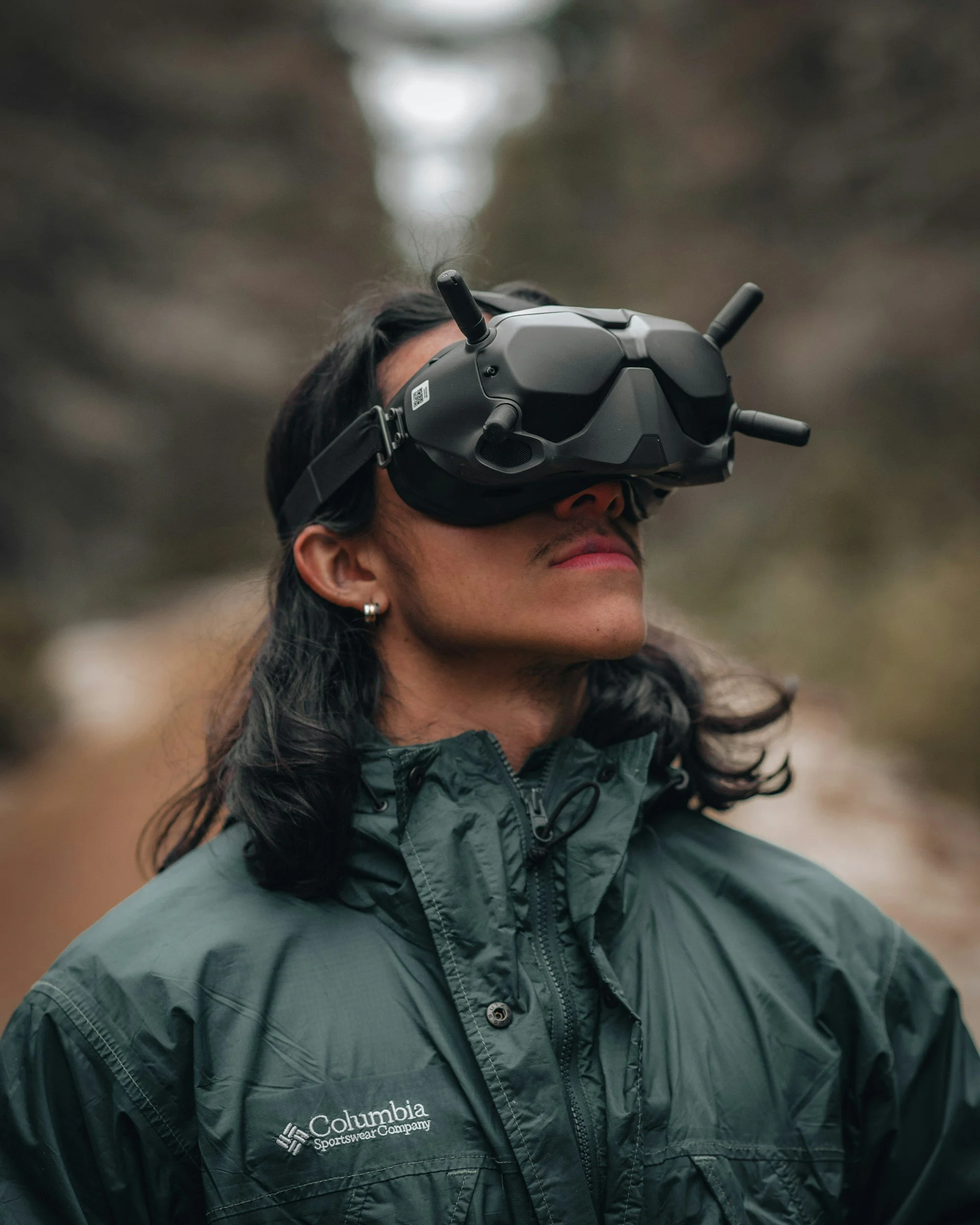 Frau mit dunklen Haaren, trägt eine schwarze Brille für Virtual Reality und eine grüne Outdoor-Jacke, steht im Freien auf einem Feldweg.