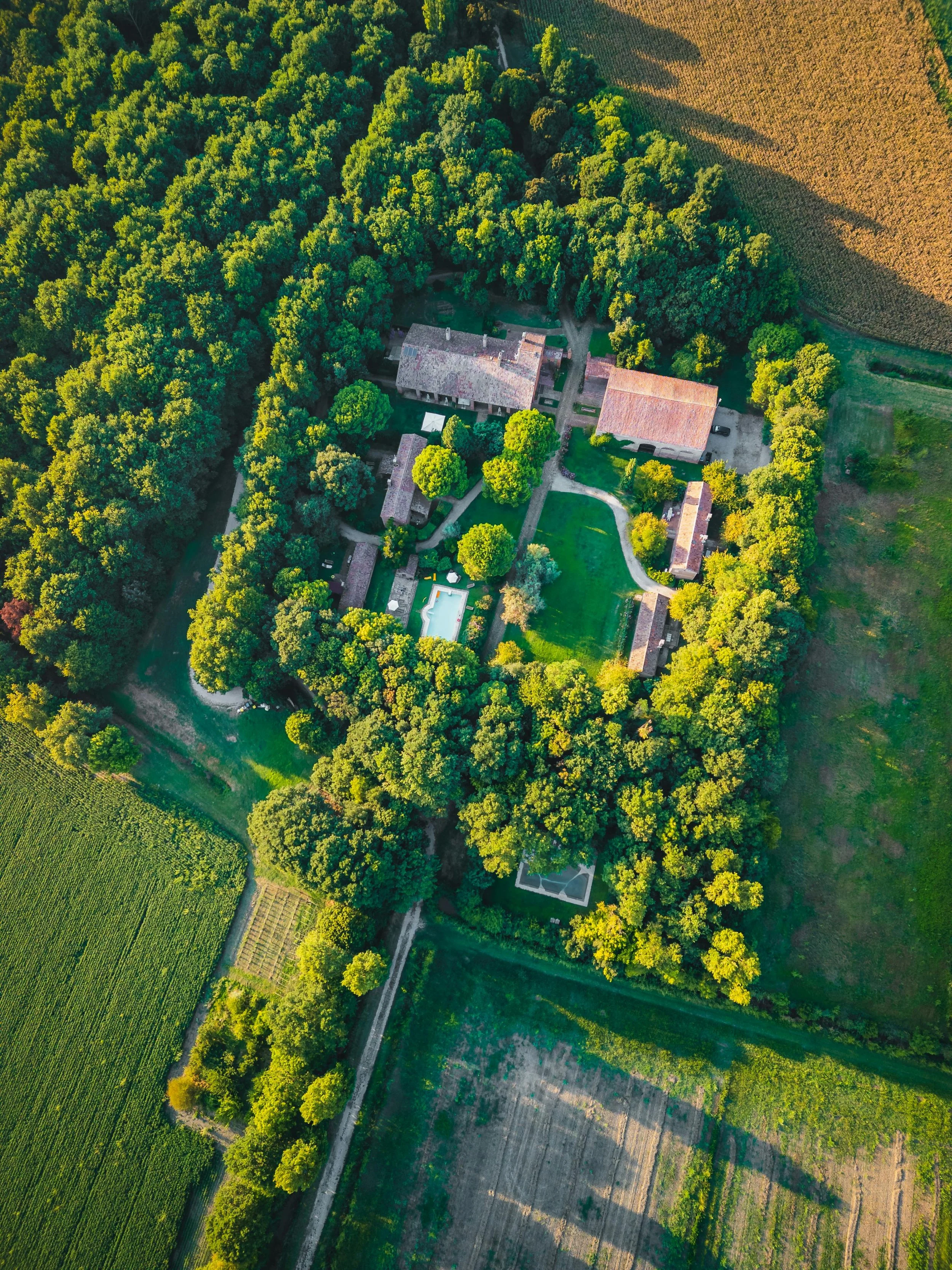 Eine Vogelperspektive auf ein großes Anwesen, umgeben von dichtem Wald und landwirtschaftlichen Feldern, mit mehreren Gebäuden, einem Pool und einem Rasen im Zentrum.