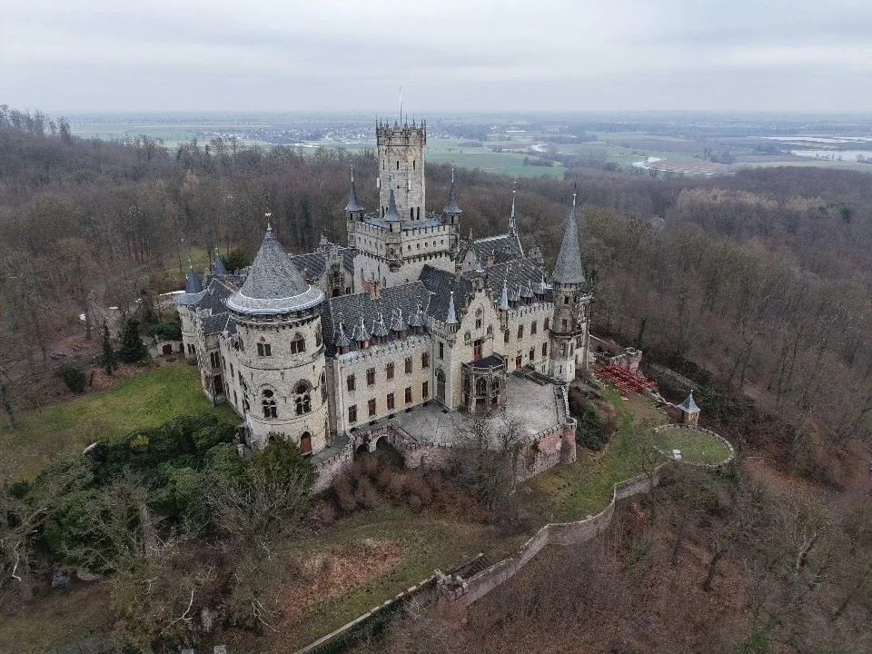 Ein mittelalterliches Schloss auf einer Anhöhe umgeben von Bäumen, mit Zinnen, Türmen und einer Mauer, bei bewölktem Himmel. Celle. Luftaufnahme Celle. Schloss Marienburg