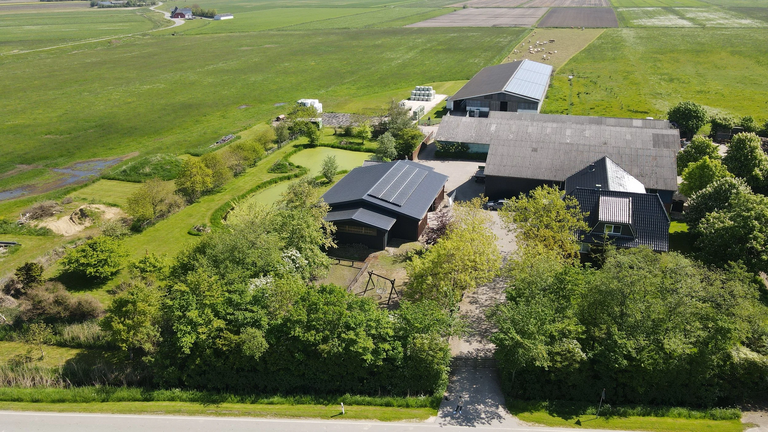 Luftaufnahme eines ländlichen Bauernhofs mit mehreren Gebäuden, umgeben von grünen Feldern und Bäumen, mit einem Spielplatz im Garten. Drohnenaufnahme Bauernhof. 