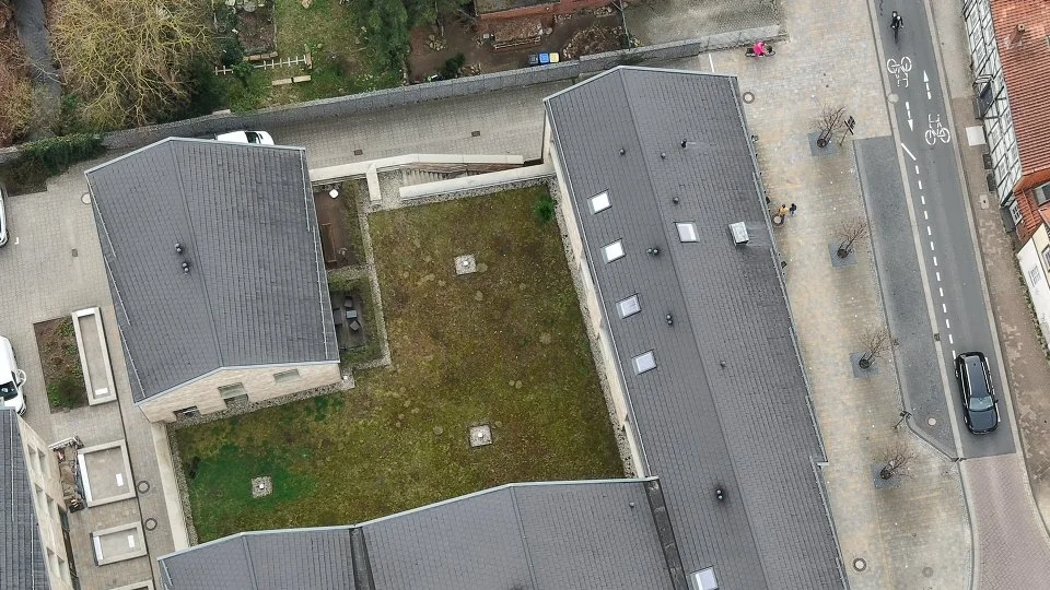 Luftaufnahme eines kleinen, quadratischen Gartens, umgeben von Gebäuden mit schwarzen Dächern, einer Straße und einem Gehweg mit Bäumen. Celle Luftaufnahme. Inspektion. Kontrolle. Luftaufnahme