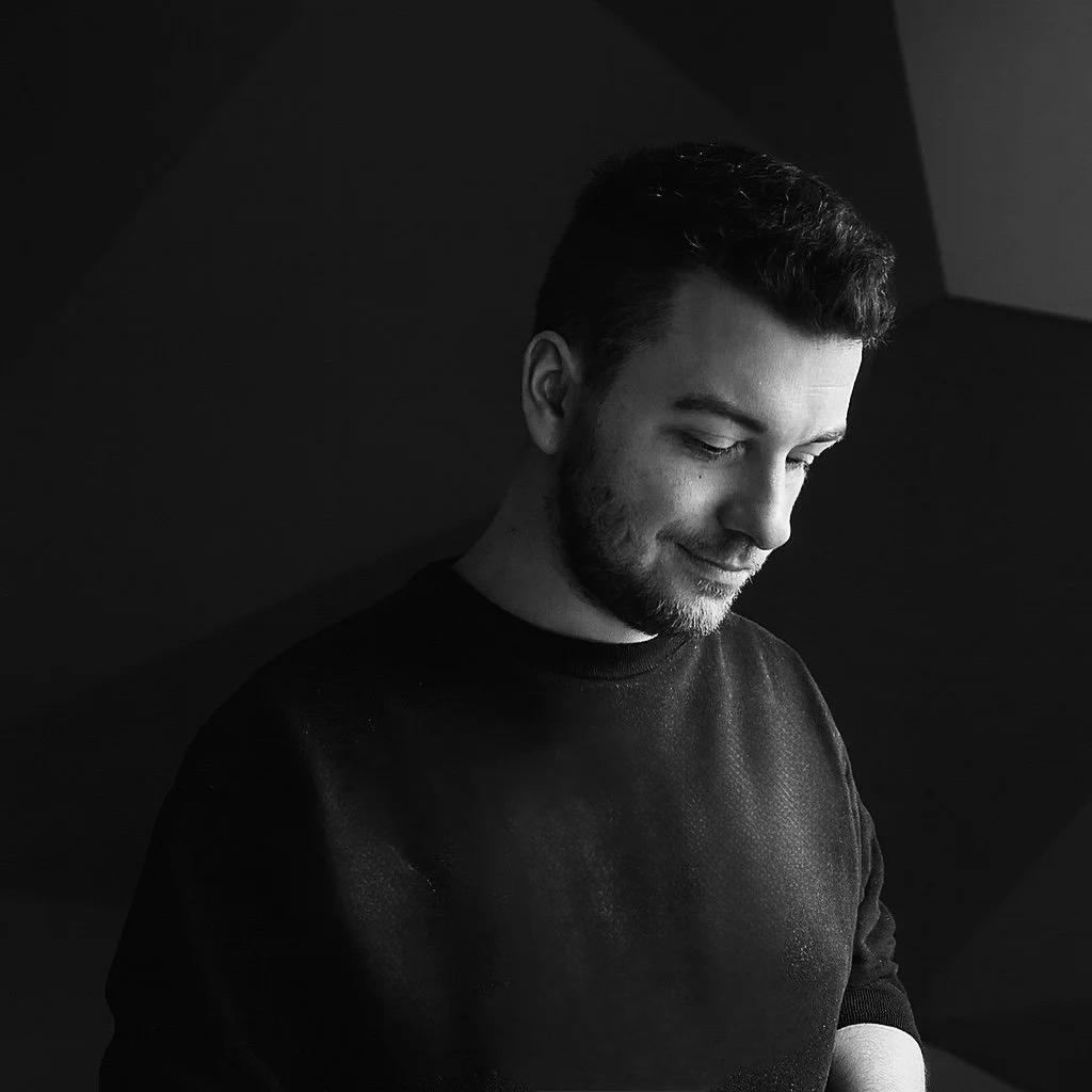 Schwarzweißfoto eines lächelnden jungen Mannes mit gepflegtem Haar und Bart, der nach unten schaut, vor dunklem Hintergrund.