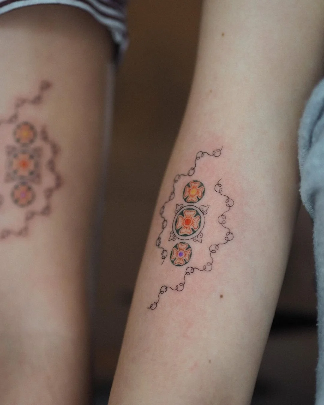 Close-up of a person's arm with a colorful floral tattoo surrounded by a black curly line design, with another blurred floral tattoo visible on the other arm.