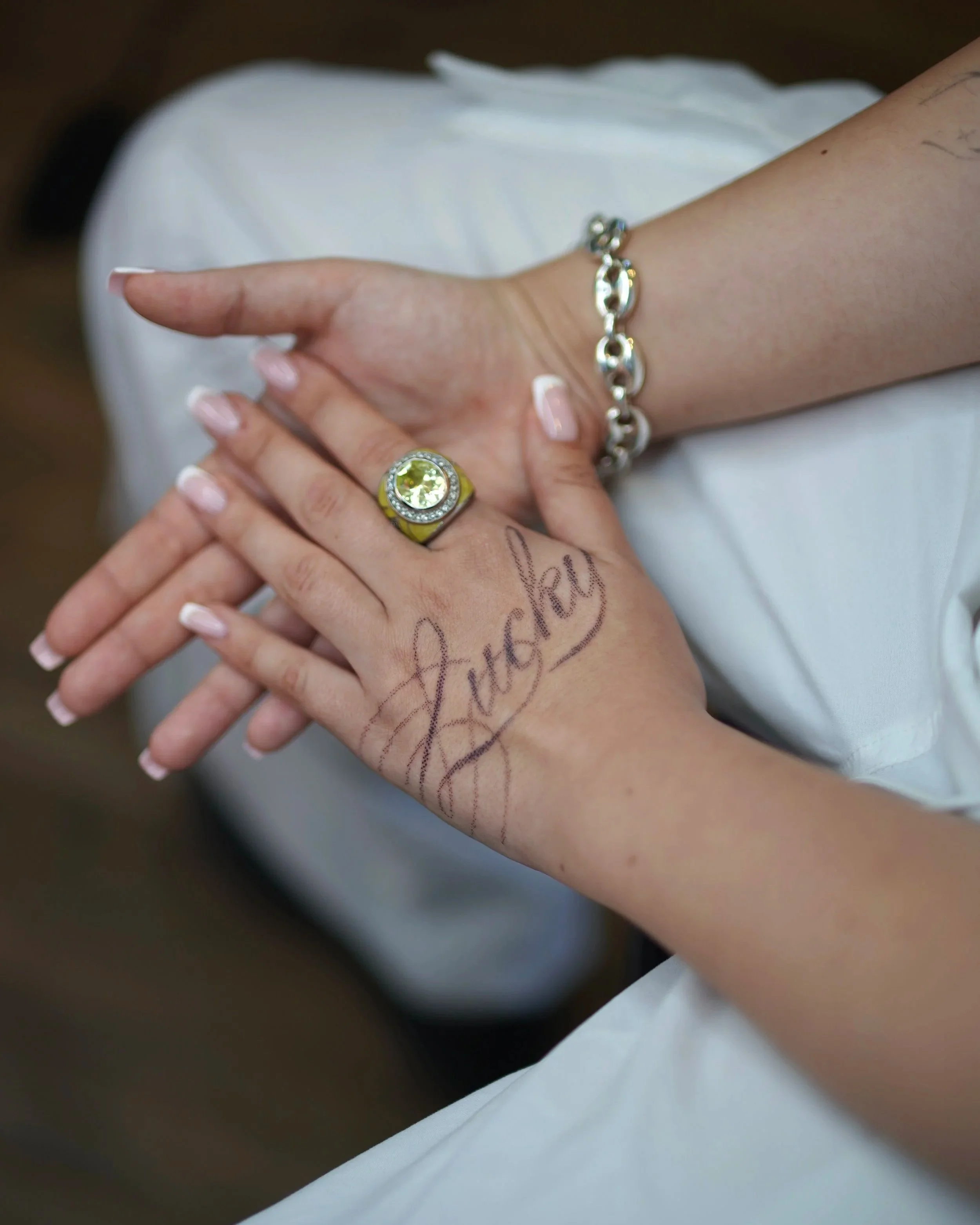 Close-up of two people holding hands, with one person wearing a yellow ring and a silver bracelet. The other person has a tattoo on their hand that reads 'family' in cursive.