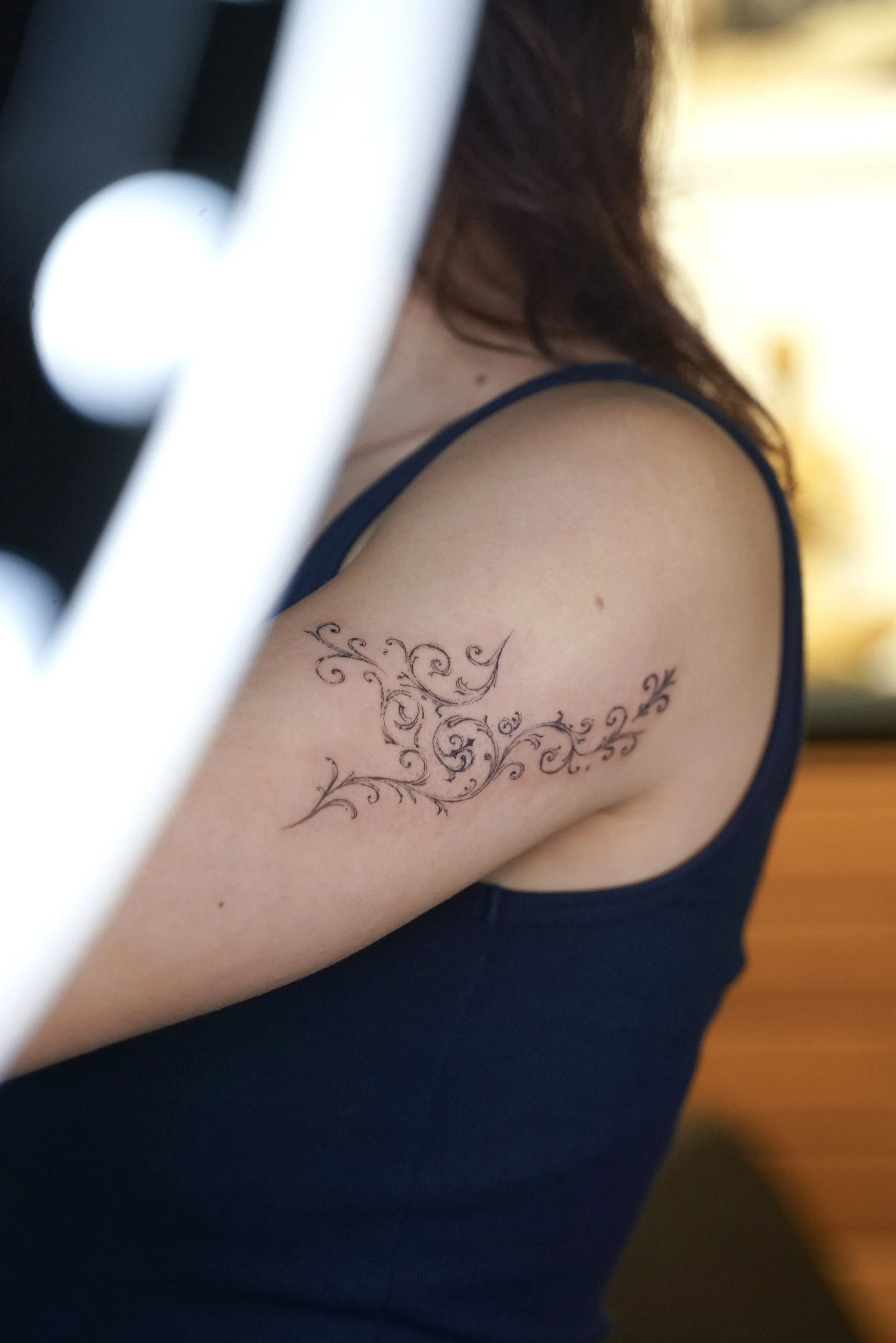 Close-up of a woman's upper arm with a black ornate tattoo of swirling floral design, wearing a dark blue sleeveless top.