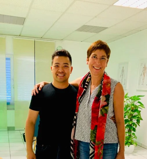 Ein junger Mann und eine ältere Frau stehen nebeneinander und lächeln, im Hintergrund sind medizinische Poster und eine Pflanze zu sehen.